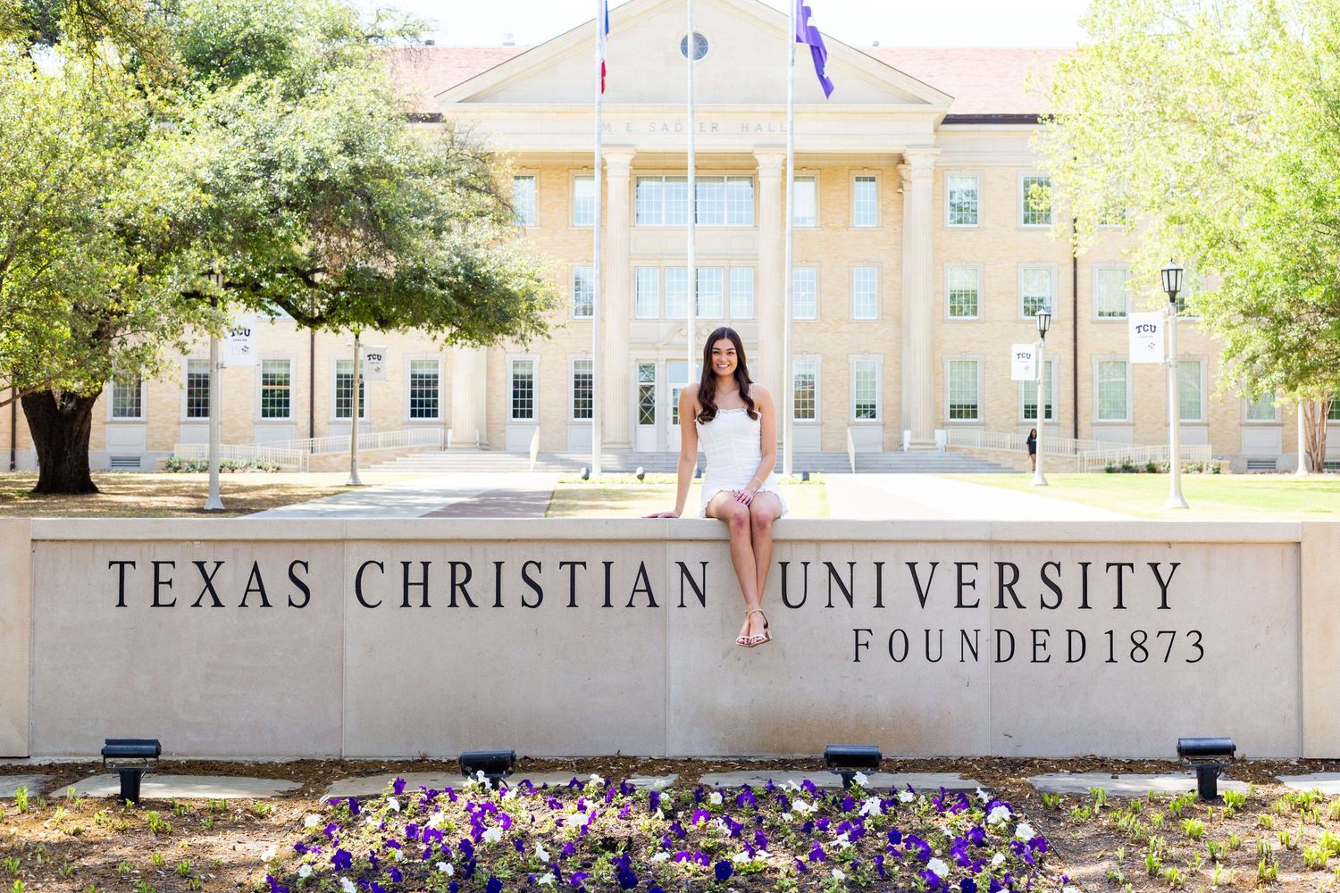 A person sits on the Texas Christian University stone entrance sign with a building and flowers in the background.