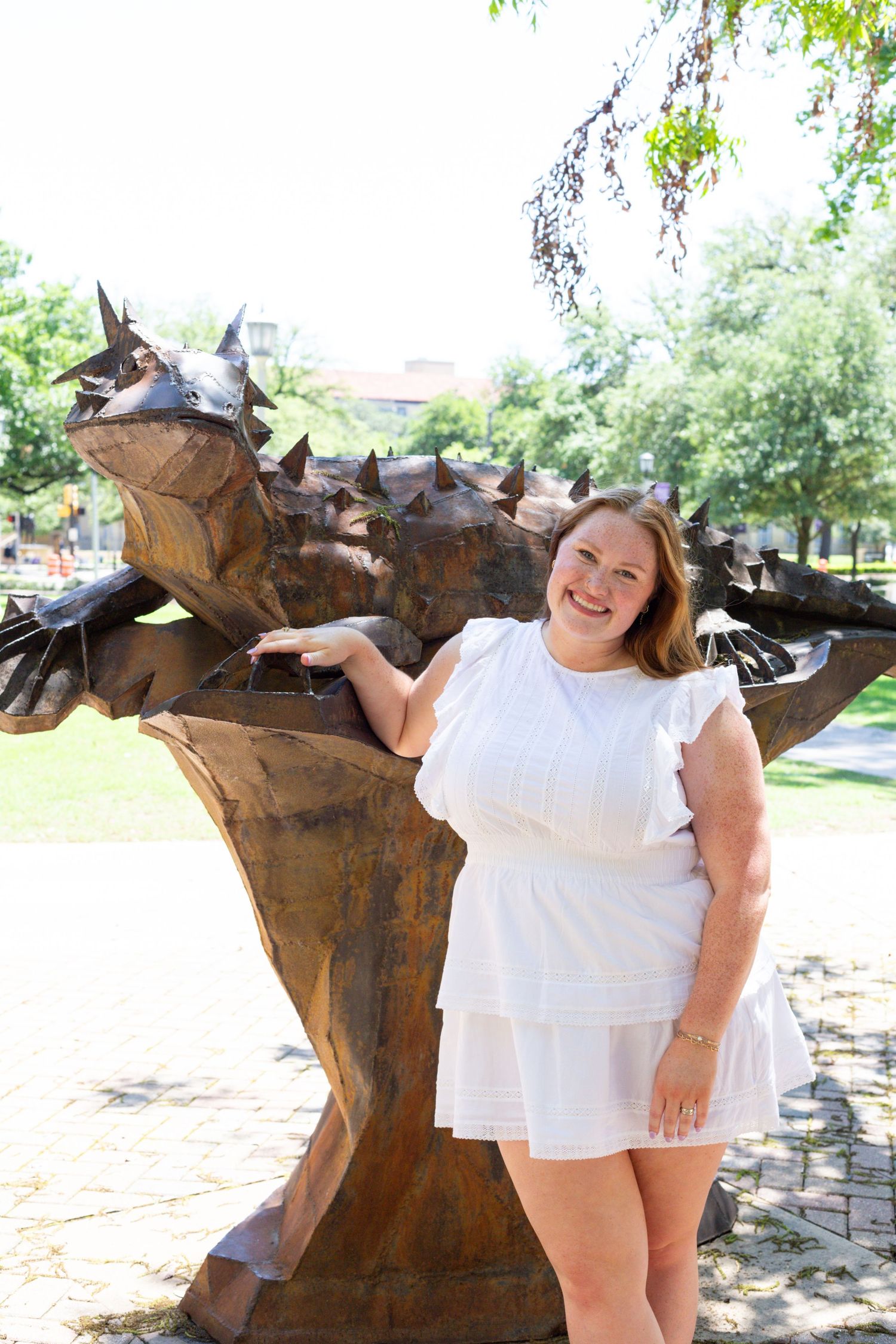 Someone in a white ruffled dress poses with a large metal dragon sculpture in an outdoor park setting.