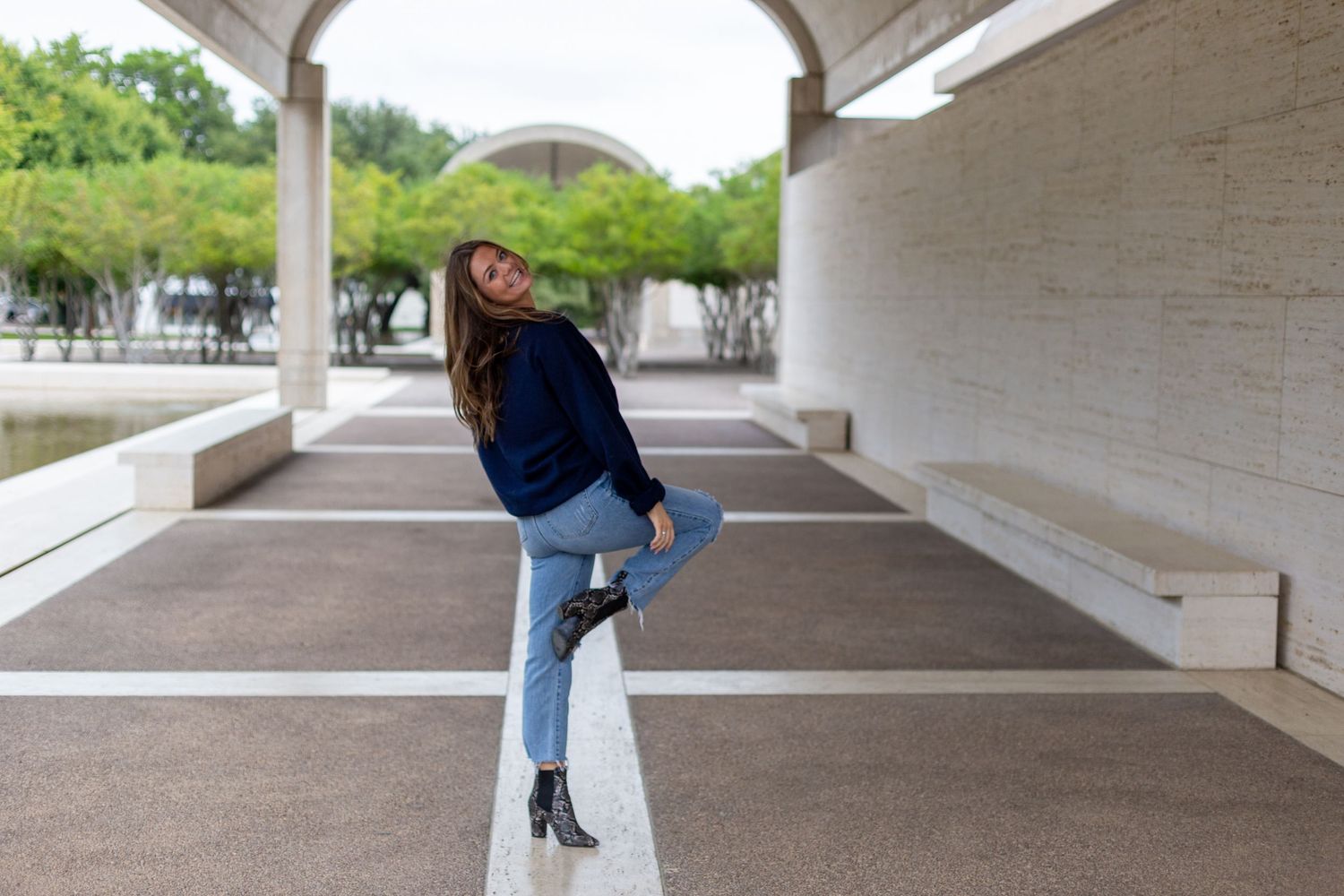 Someone in a blue sweater and jeans poses playfully in an outdoor covered walkway with white columns and arches.