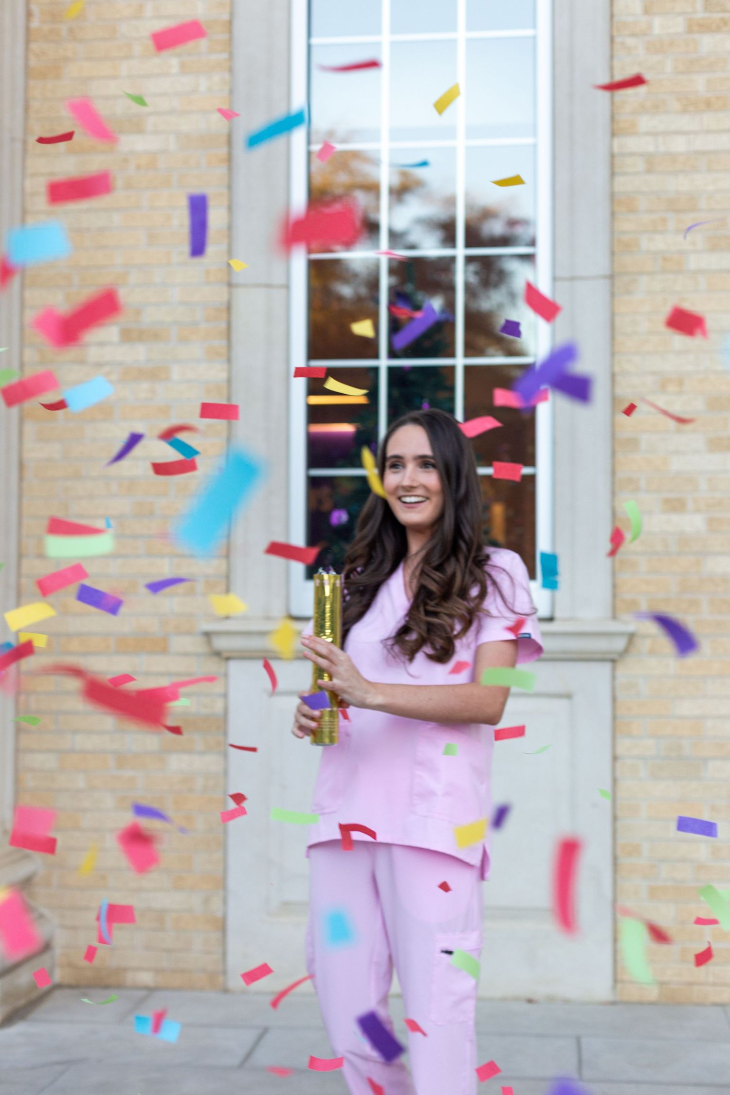 A graduate in pink scrubs celebrates with colorful confetti falling outside a brick building.