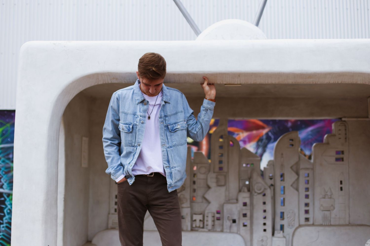Casual street style outfit featuring light denim jacket and dark pants against white architectural backdrop with hanging flags.