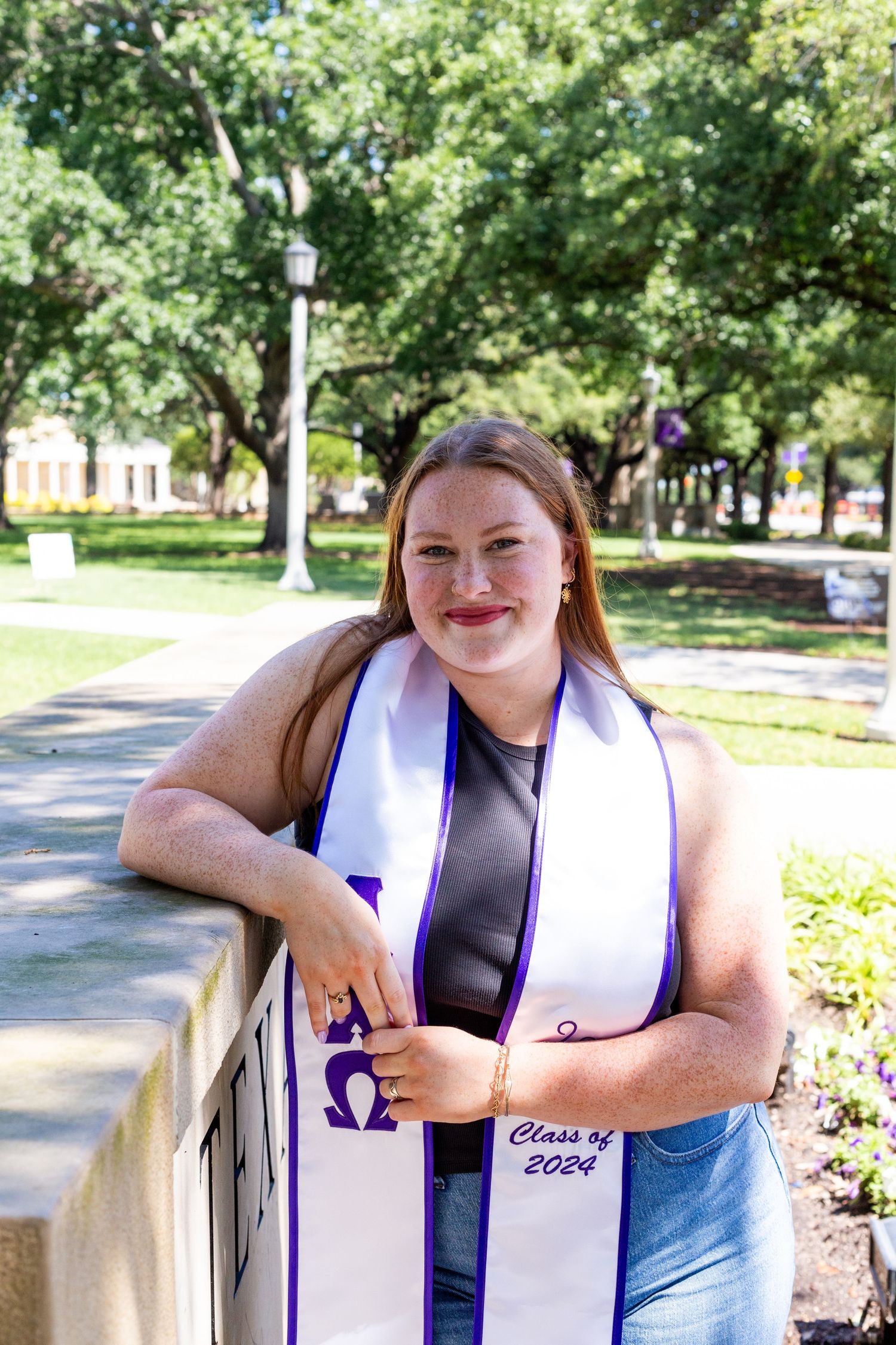A person wearing a graduation stole with Greek letters stands smiling in a park setting during a sunny day.