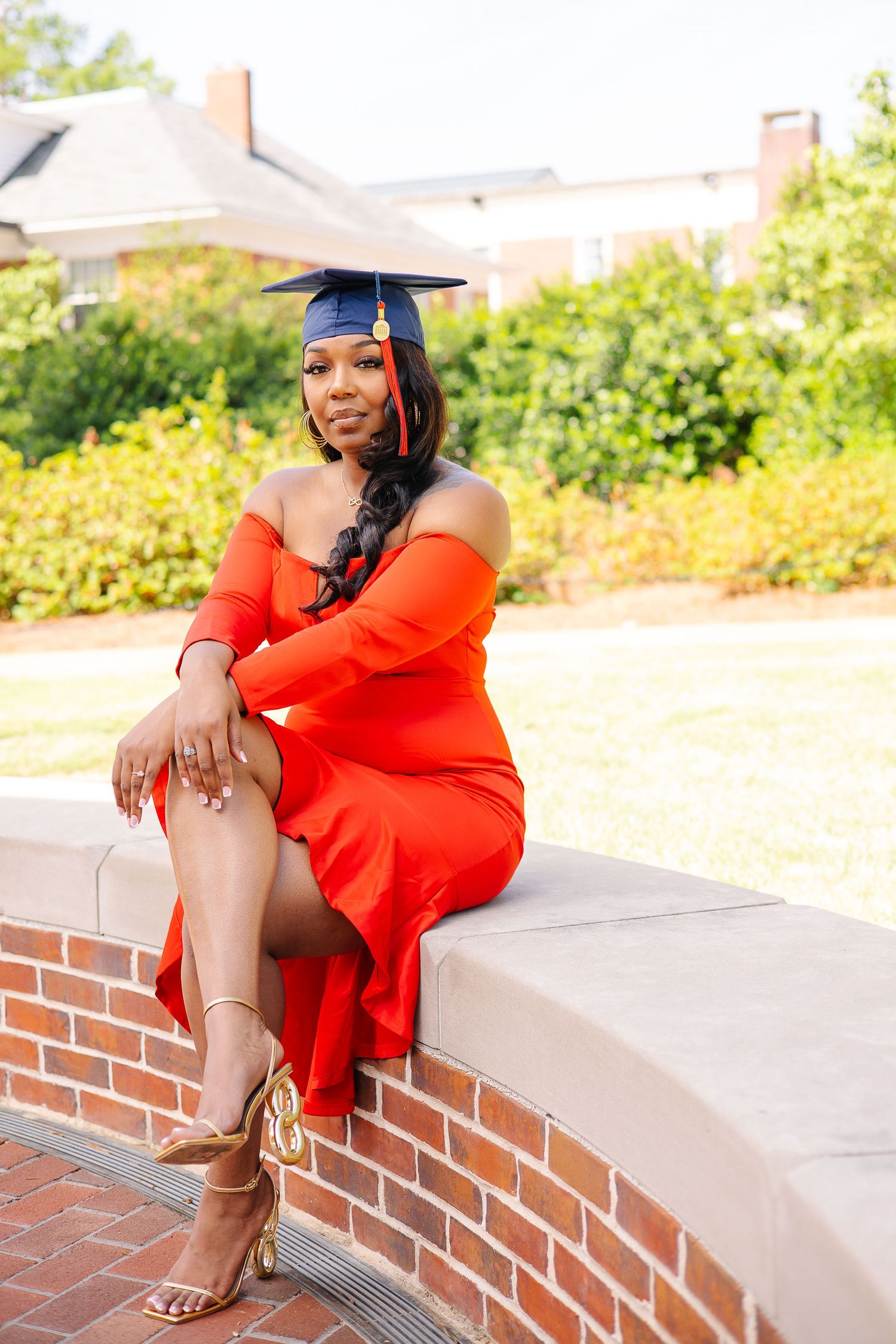 A woman in a red off-shoulder dress and graduation cap poses on a brick wall outdoors for her graduation photos.