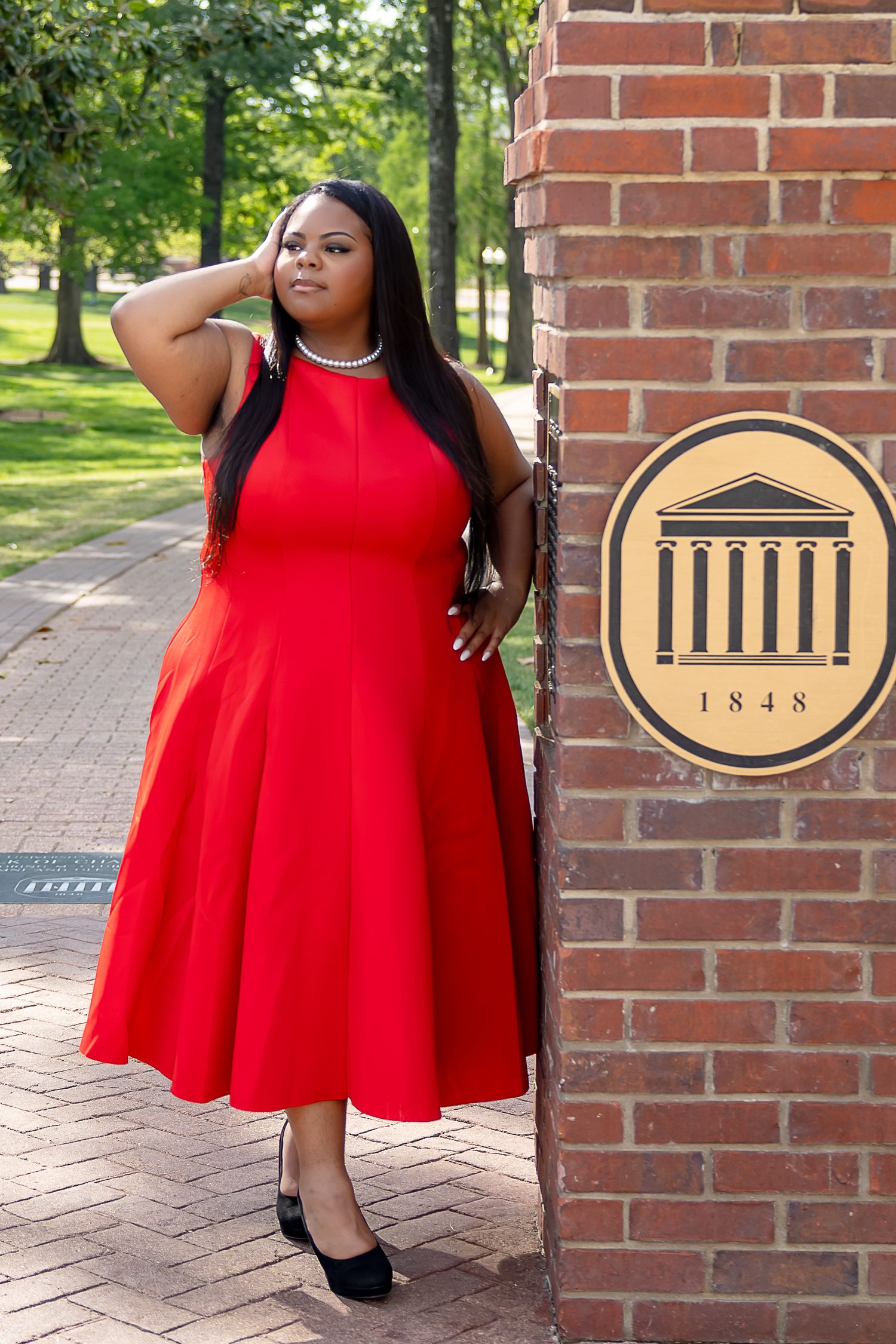 Woman in red dress posing by brick wall with 1848 university seal on a tree-lined campus path.