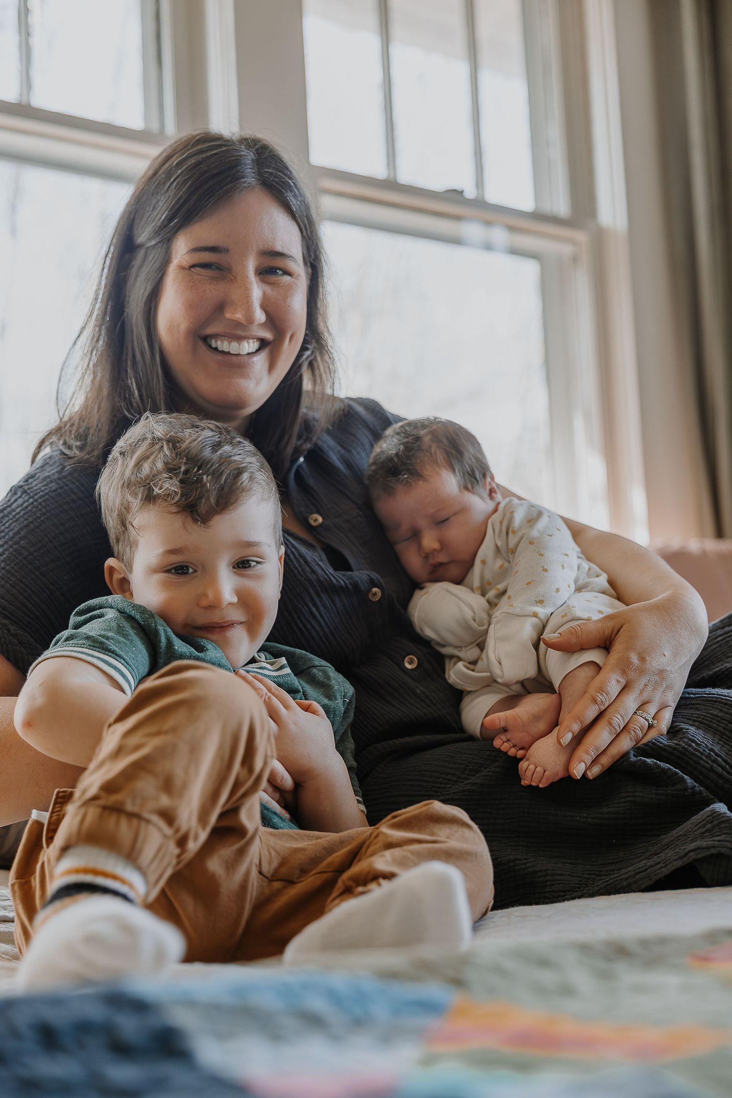 A tender moment of family bonding shows parent and child cuddling together on a cozy surface near sunlit windows.