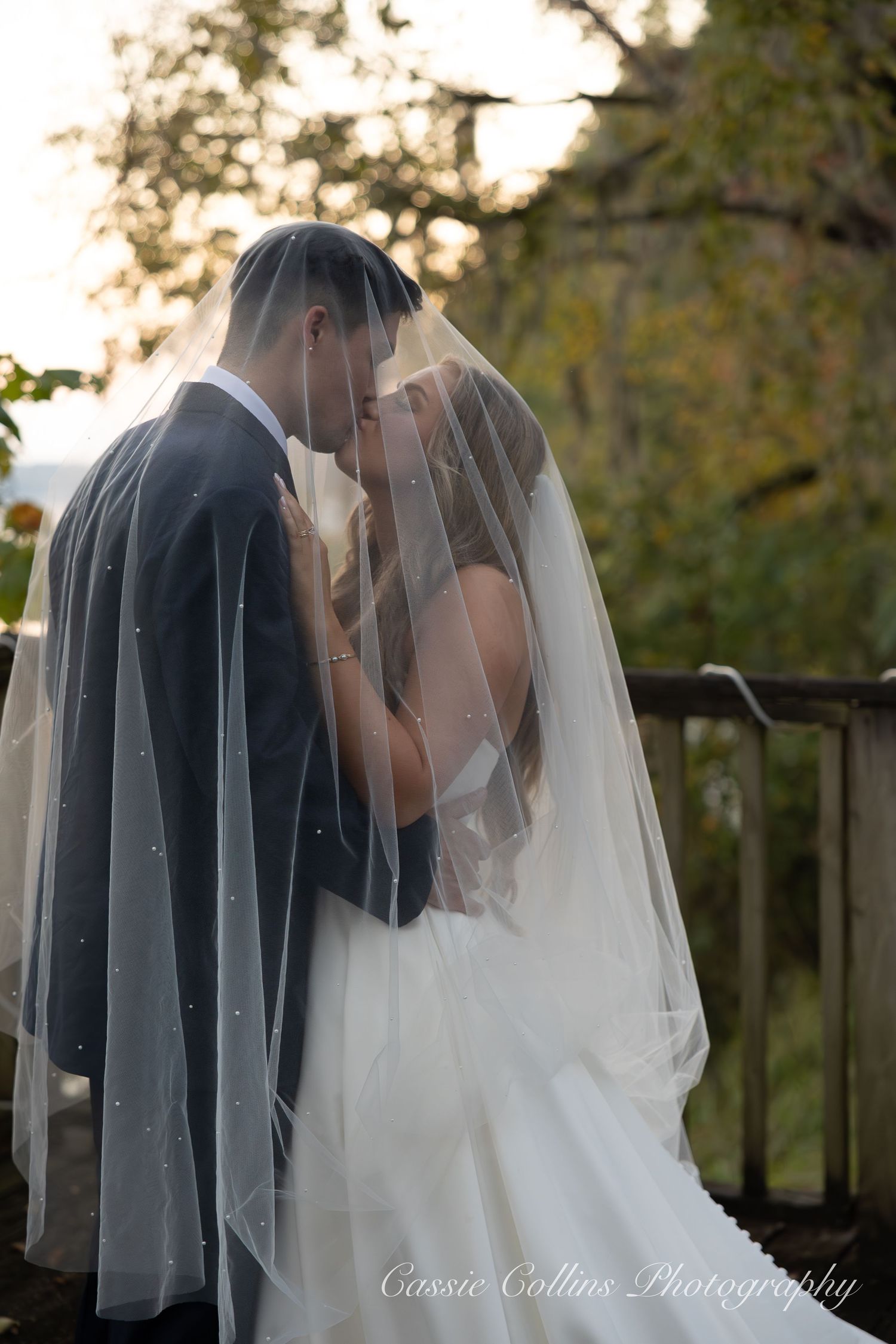 Romantic wedding couple shares intimate moment at sunset under trees with bride's veil flowing in soft light.