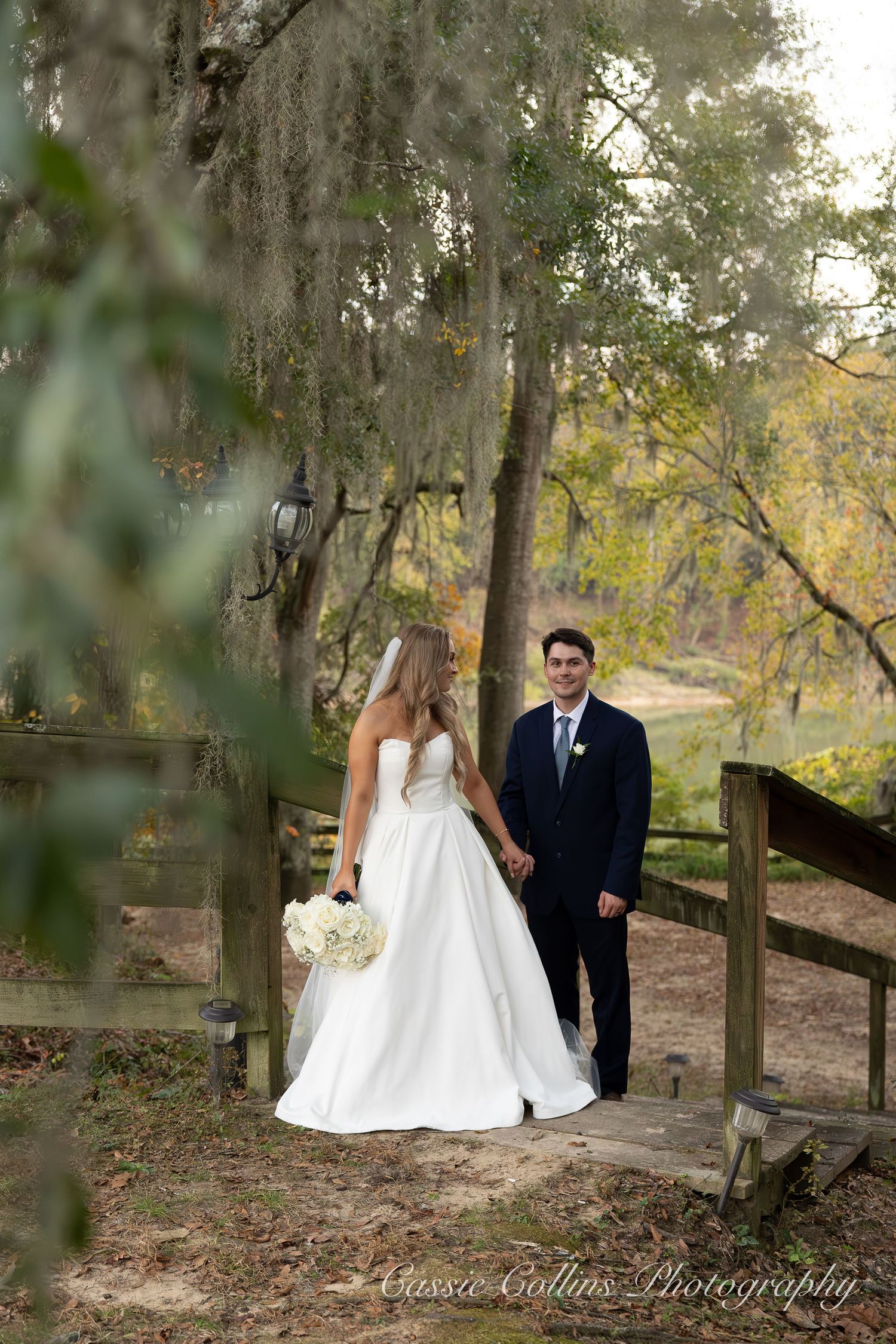 Bride in white gown and groom in navy suit hold hands on rustic wooden steps surrounded by Spanish moss-draped trees.