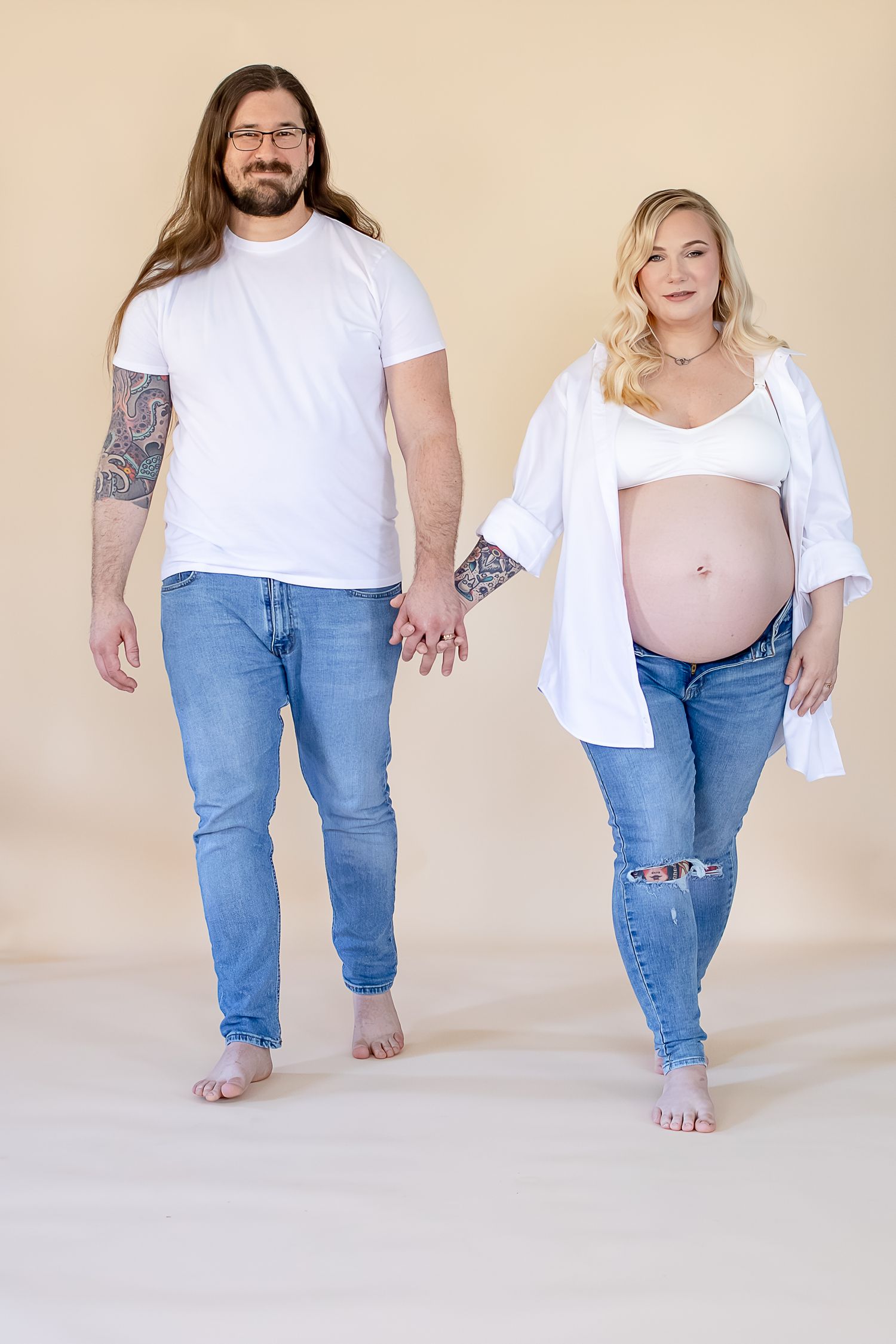 A couple in casual denim and white tops pose barefoot together, with the woman showing her pregnant belly.