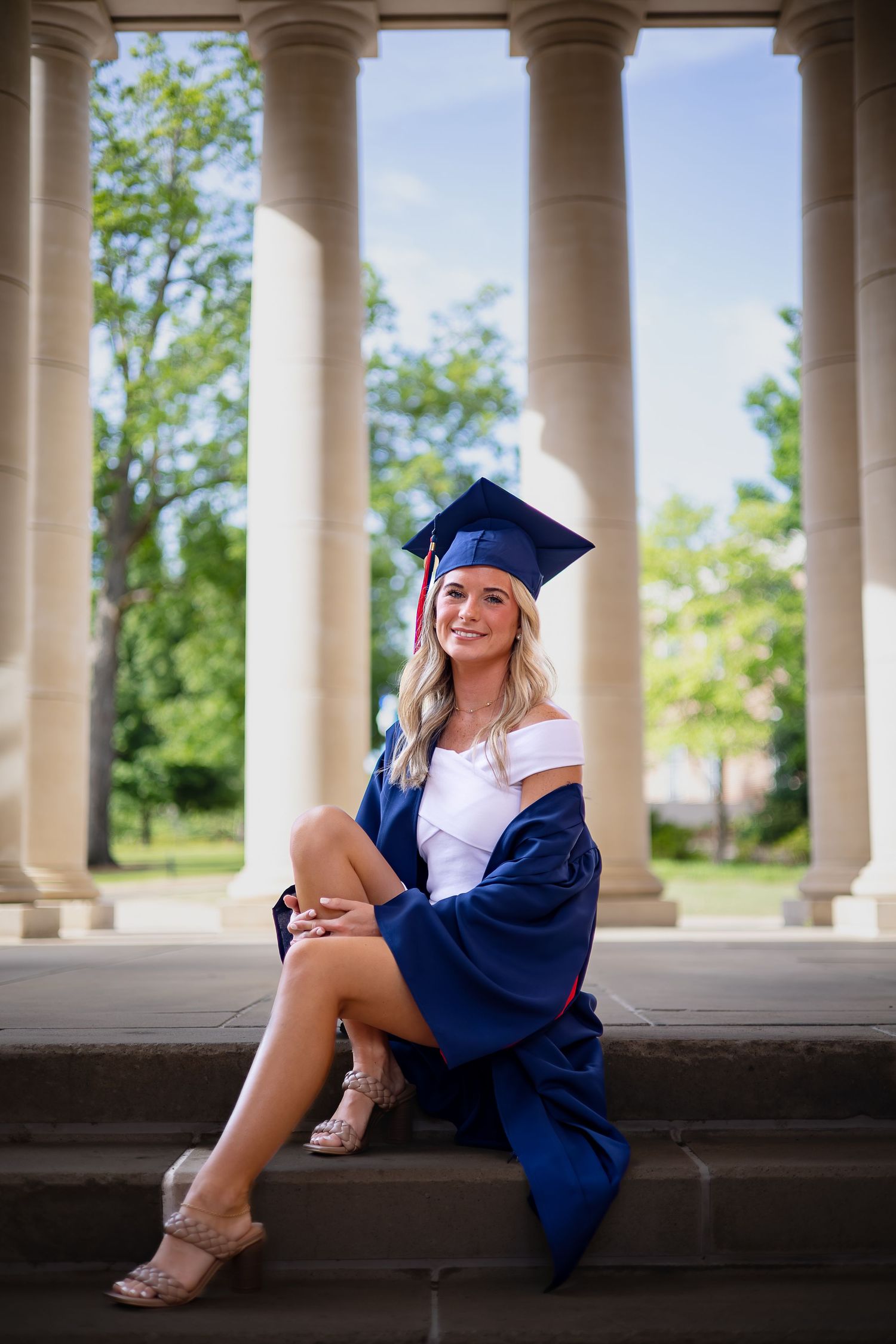 Smiling graduate in blue cap and gown sitting on steps between white columns.