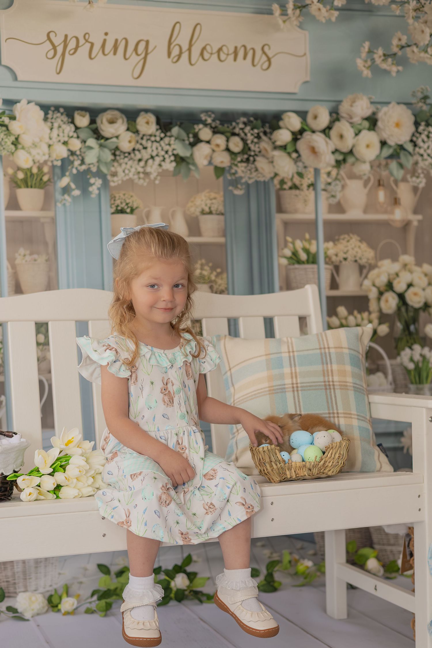 Spring themed photo session with white floral decorations, rustic wooden bench, and soft blue wall background.