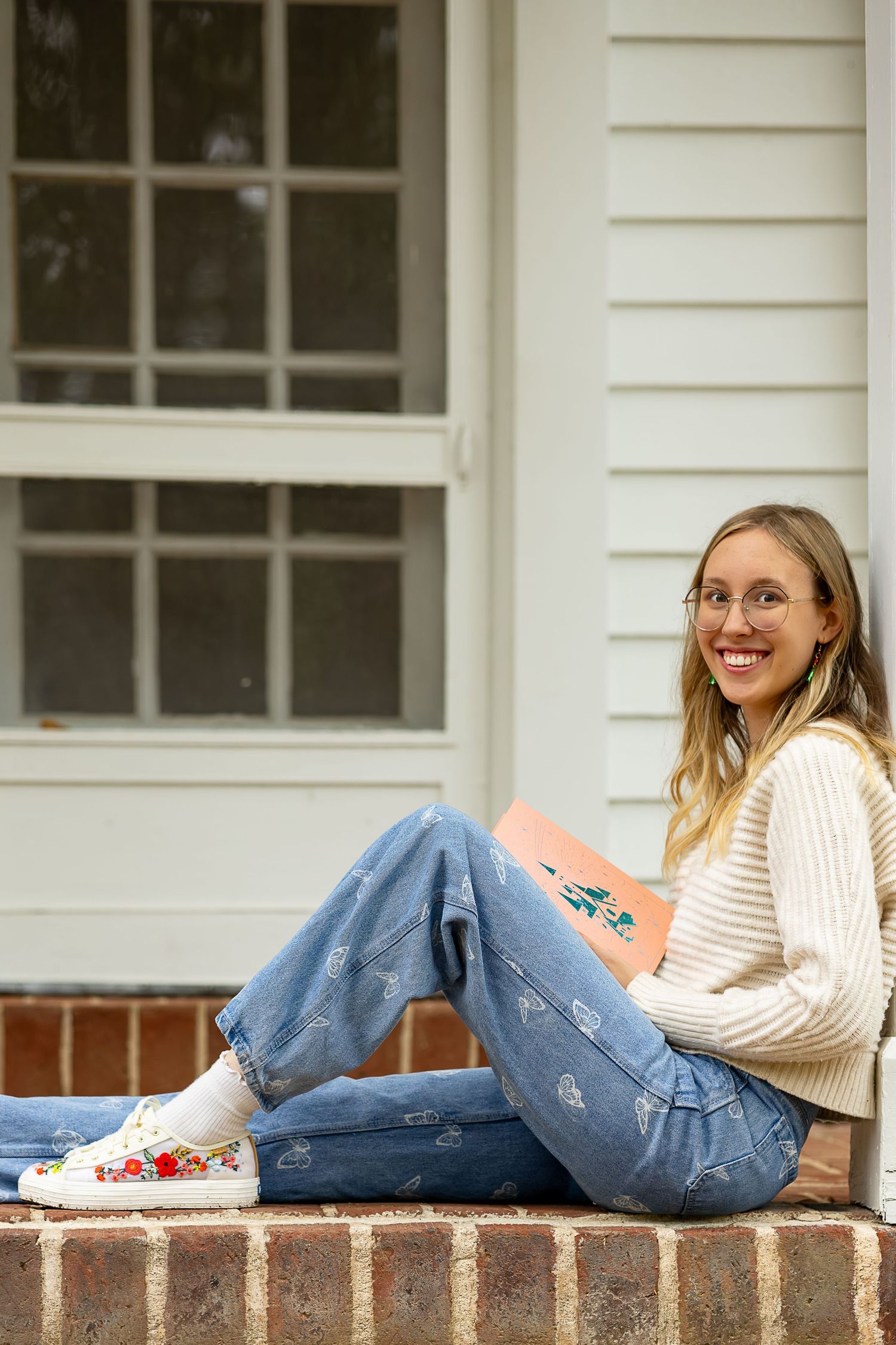 Young woman in jeans and floral sneakers sitting on brick steps, smiling while holding a pink book outdoors.