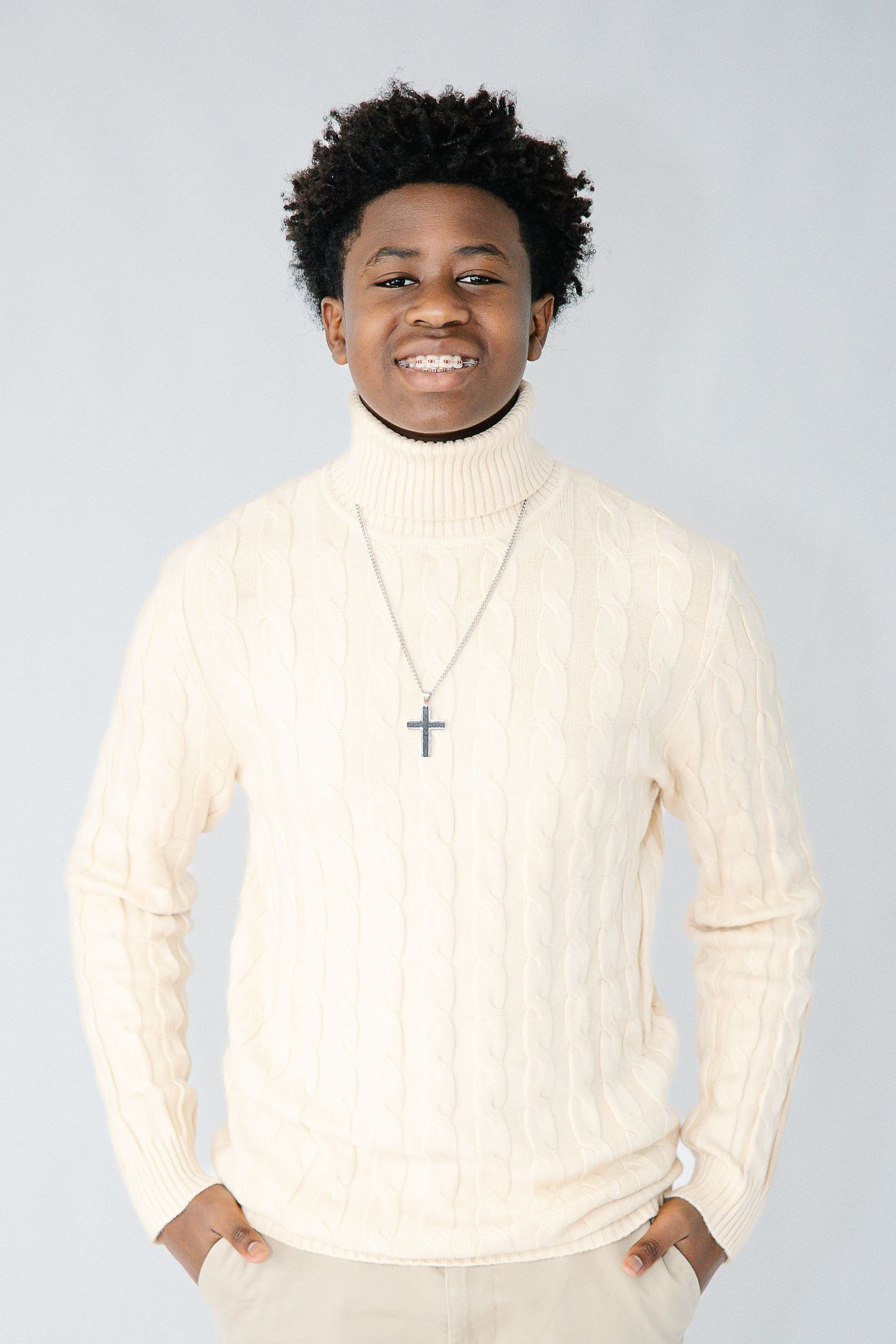 A person wearing a cream cable knit turtleneck sweater and silver cross necklace smiles against a light background.