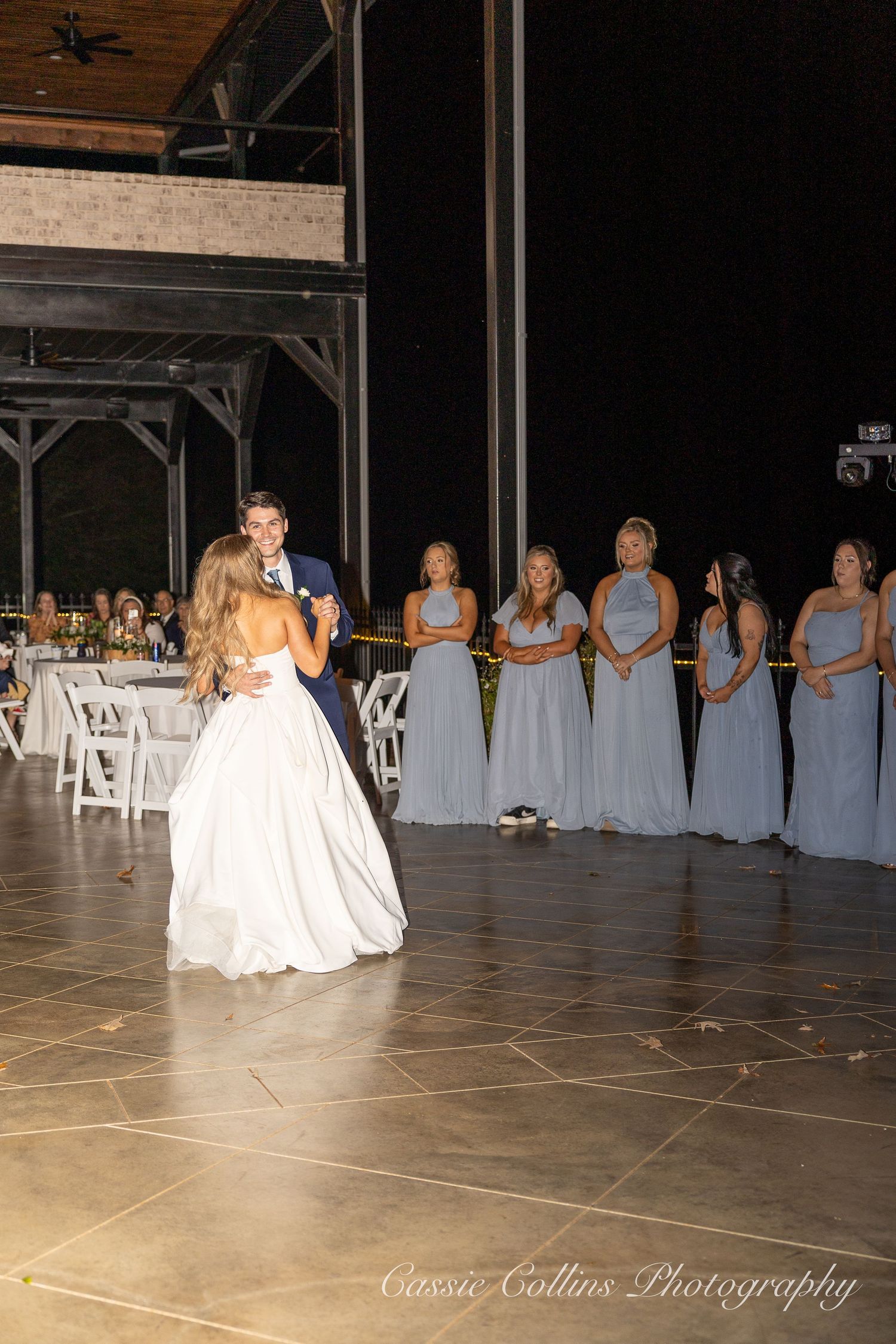 A couple shares their first dance at a wedding reception while bridesmaids in light blue dresses watch from the sidelines.