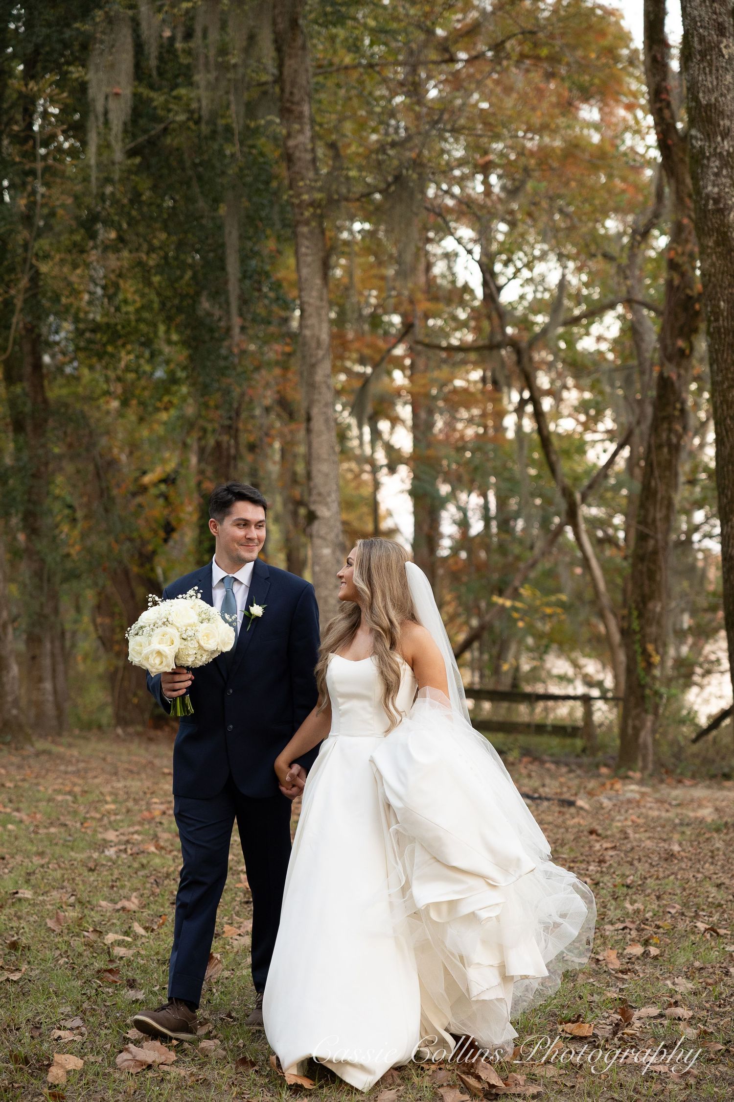 Bride and groom holding hands in a wooded autumn setting, bride in white gown with veil, groom in navy suit.