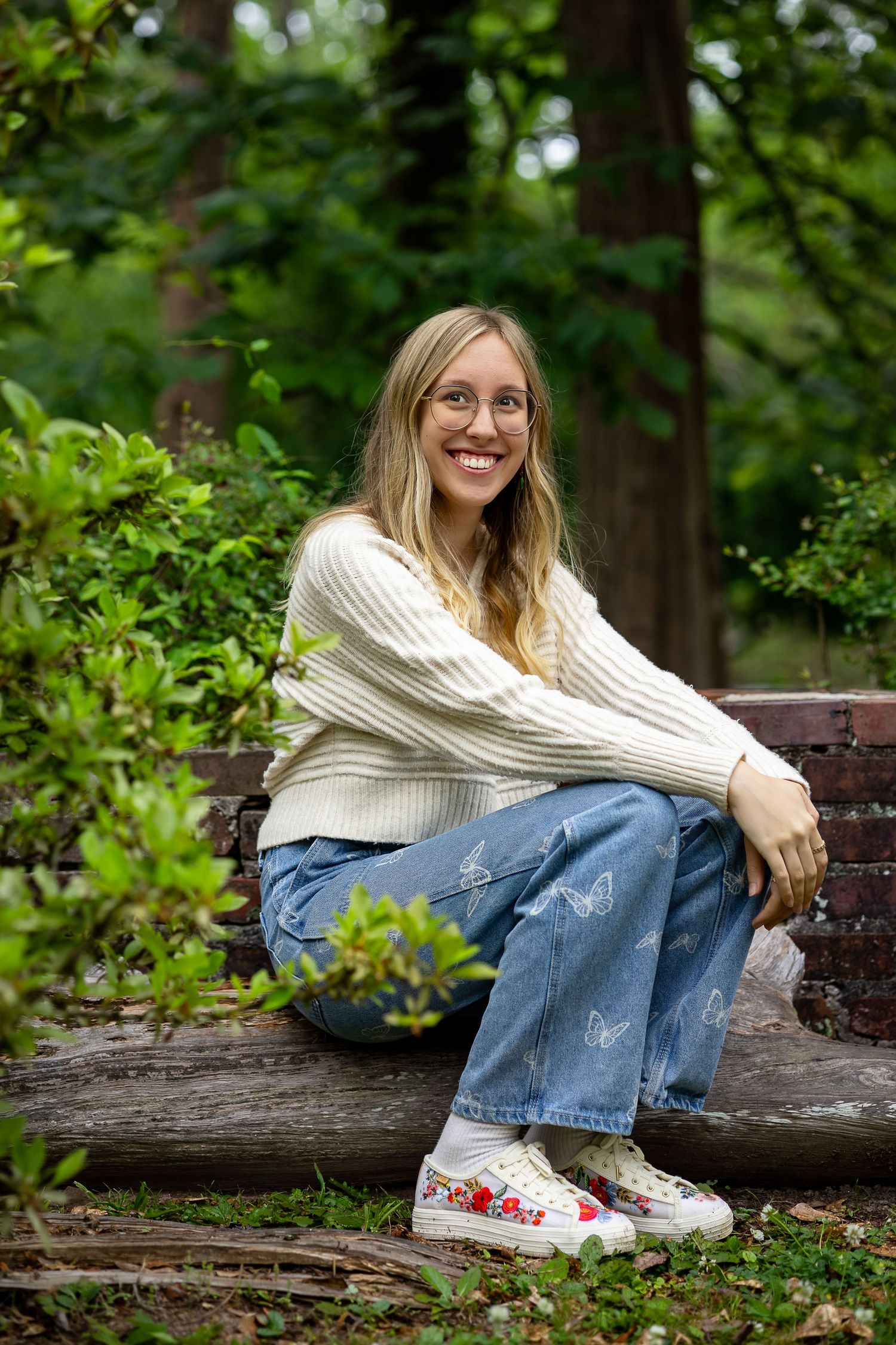 Happy blonde woman wearing glasses and jeans sitting on a wooden bench surrounded by lush green garden foliage.
