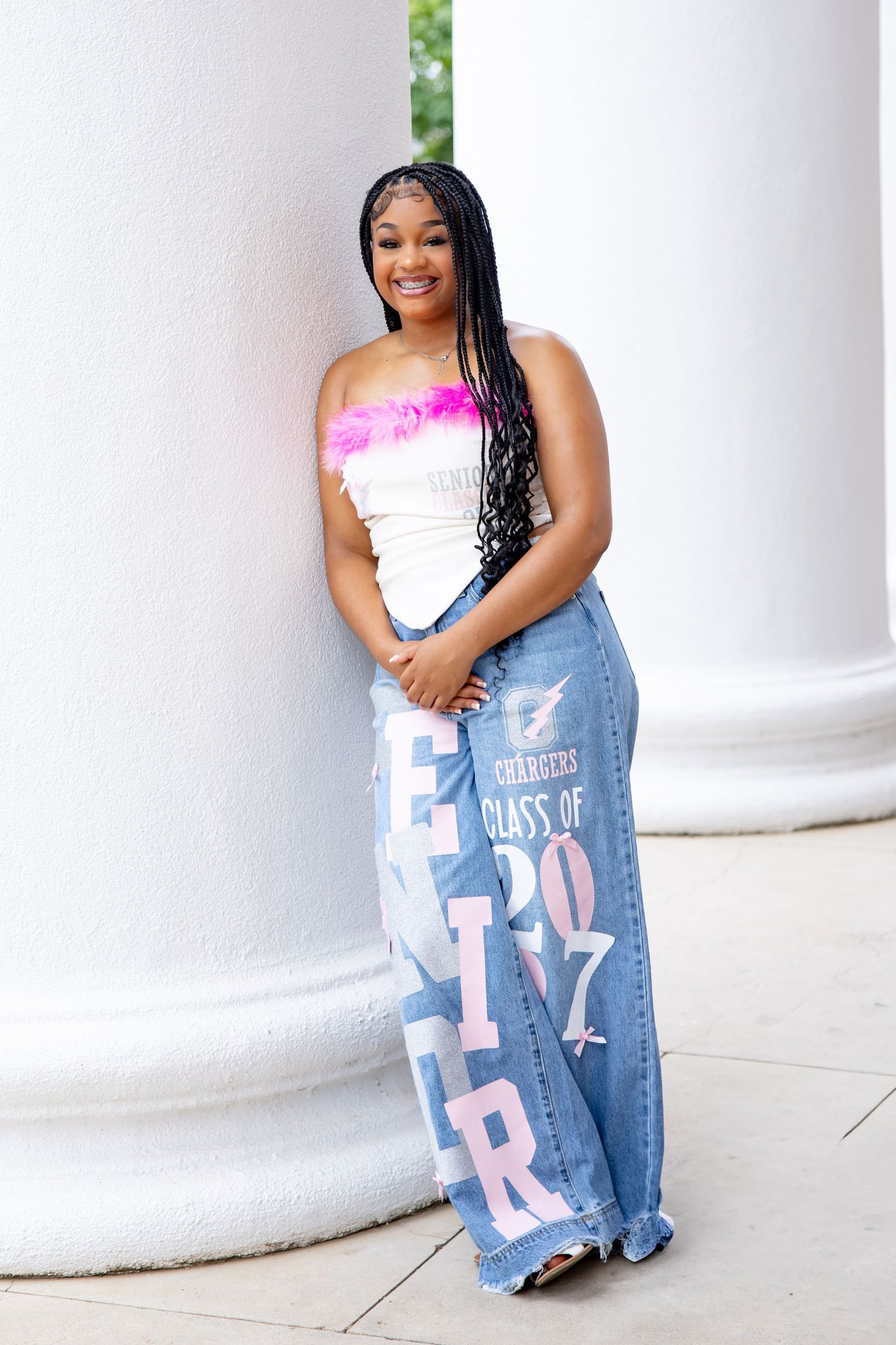 Young woman in white feather tube top and Class of 2024 wide-leg jeans smiling by white columns.