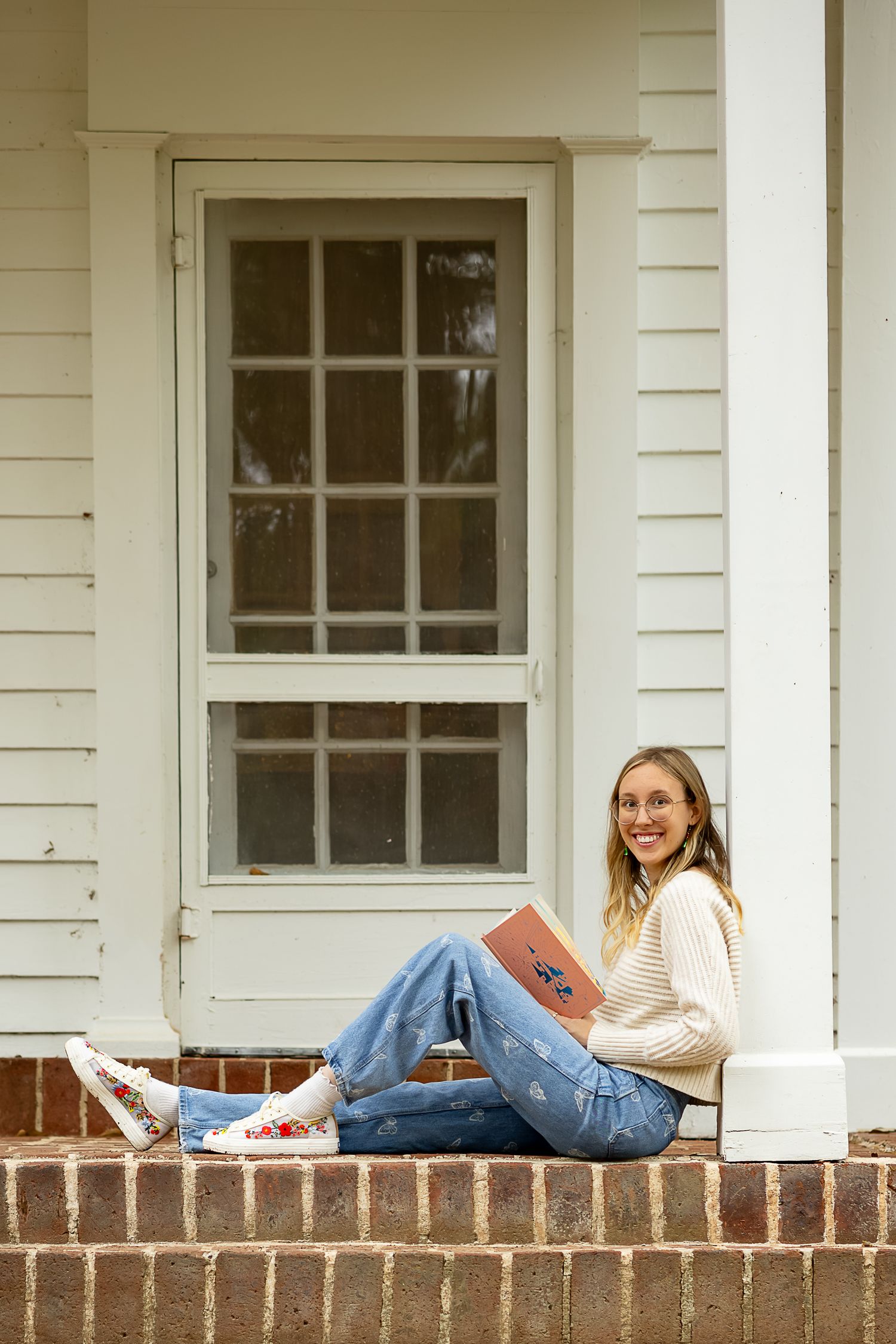 Young woman in jeans and sweater sitting on brick porch steps, smiling while holding a book.