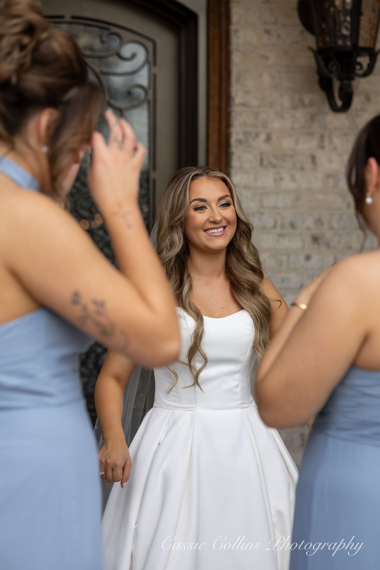 A bride in a white dress shares a candid moment with bridesmaids in light blue dresses at a rustic brick venue.