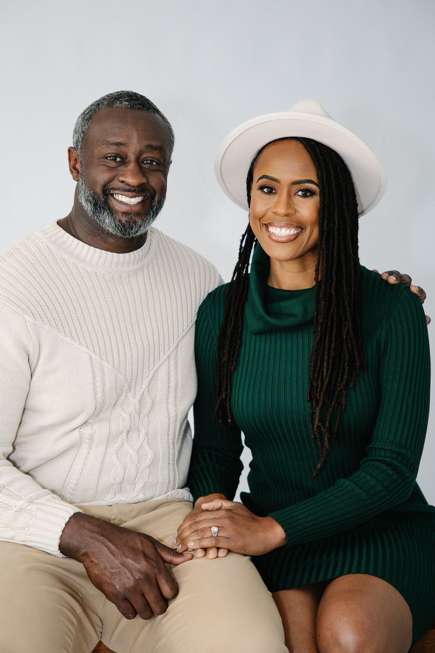 A couple wearing white and green sweaters sit together on a white couch in a minimalist studio setting.