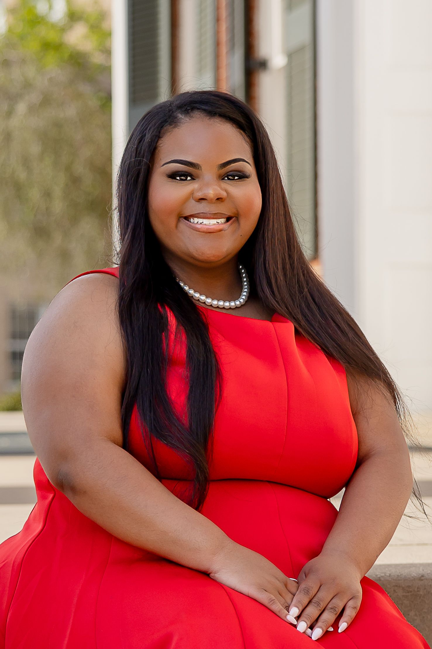 Smiling woman in red sleeveless dress and pearl necklace sitting outdoors in a professional portrait photo.