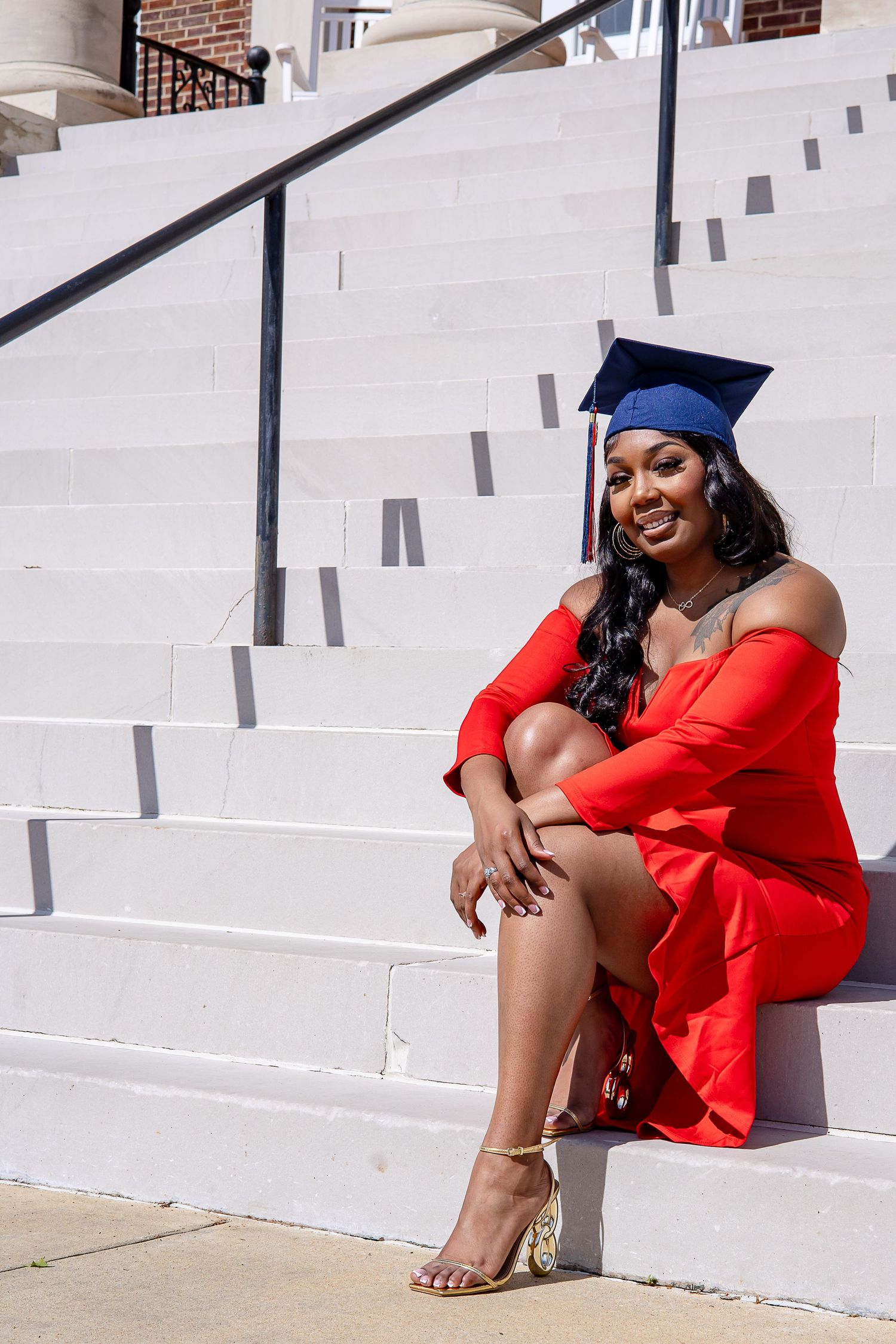 Smiling graduate in red off-shoulder dress and blue cap sits on white steps celebrating her achievement.