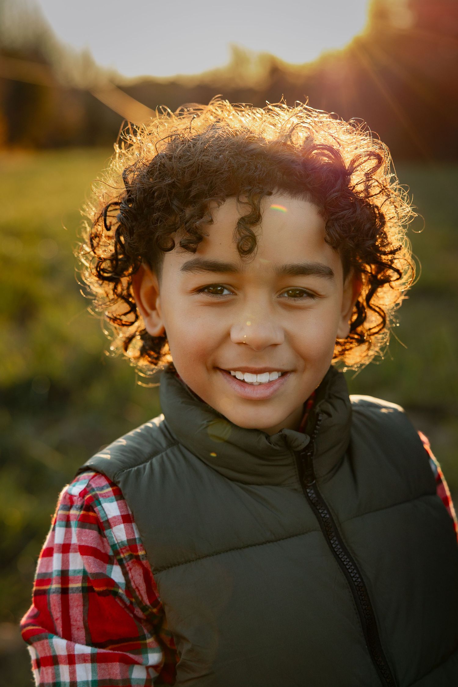 Child with curly hair wearing black vest and plaid shirt smiles warmly during golden hour photoshoot.