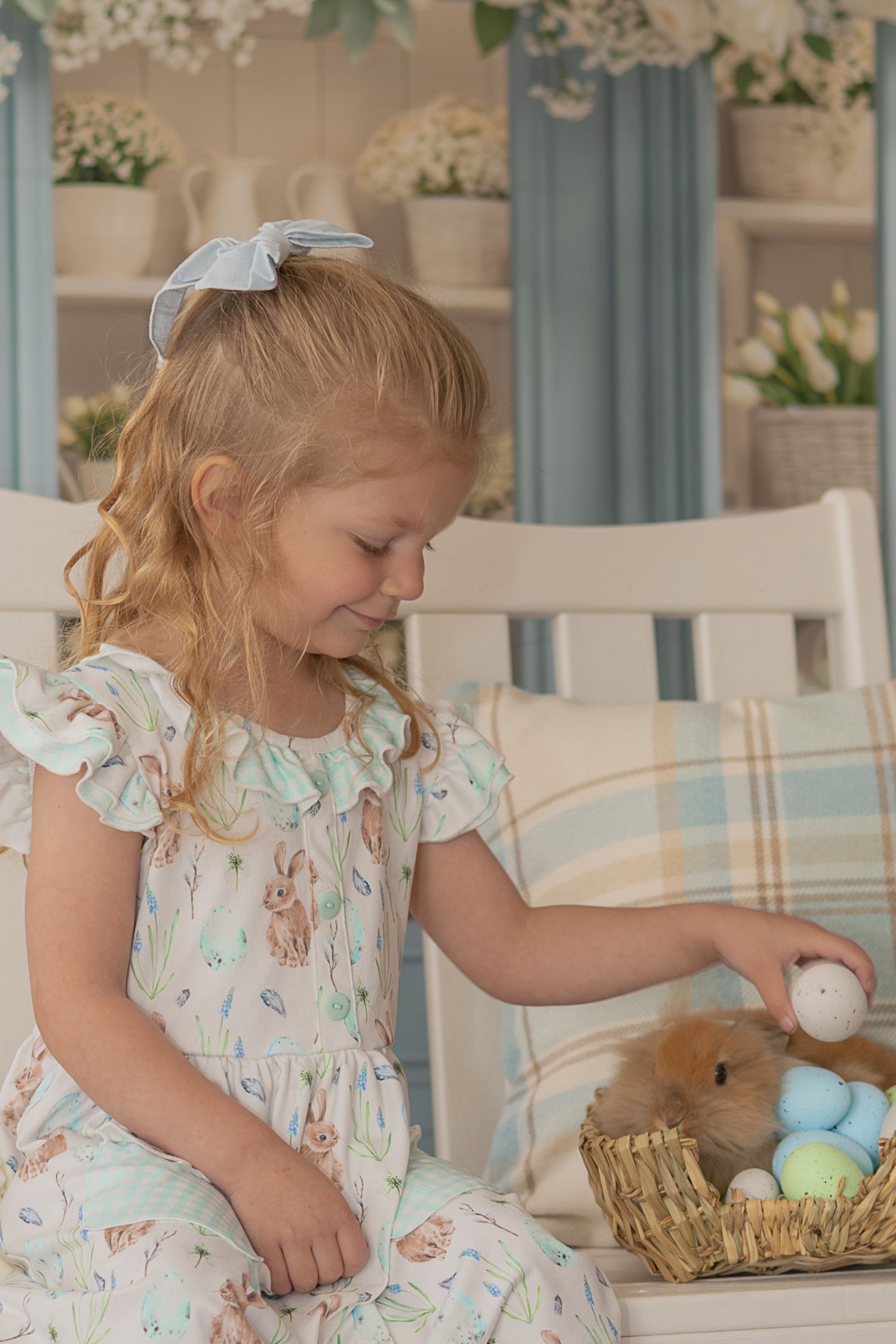 Young child in floral dress reaching into Easter basket filled with colorful eggs on white table.