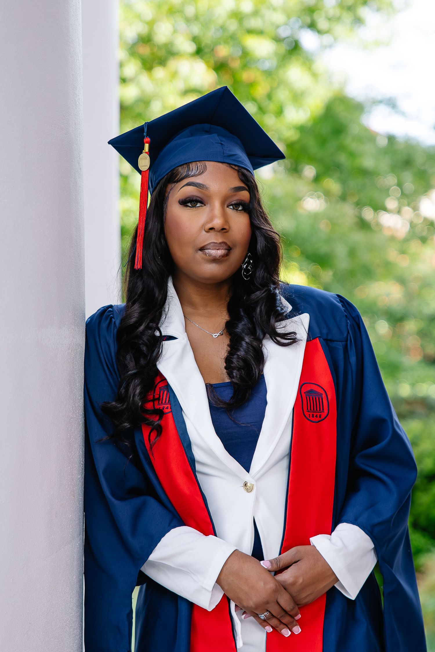 Young woman in navy blue graduation cap and gown with red sash poses confidently outdoors near white column.
