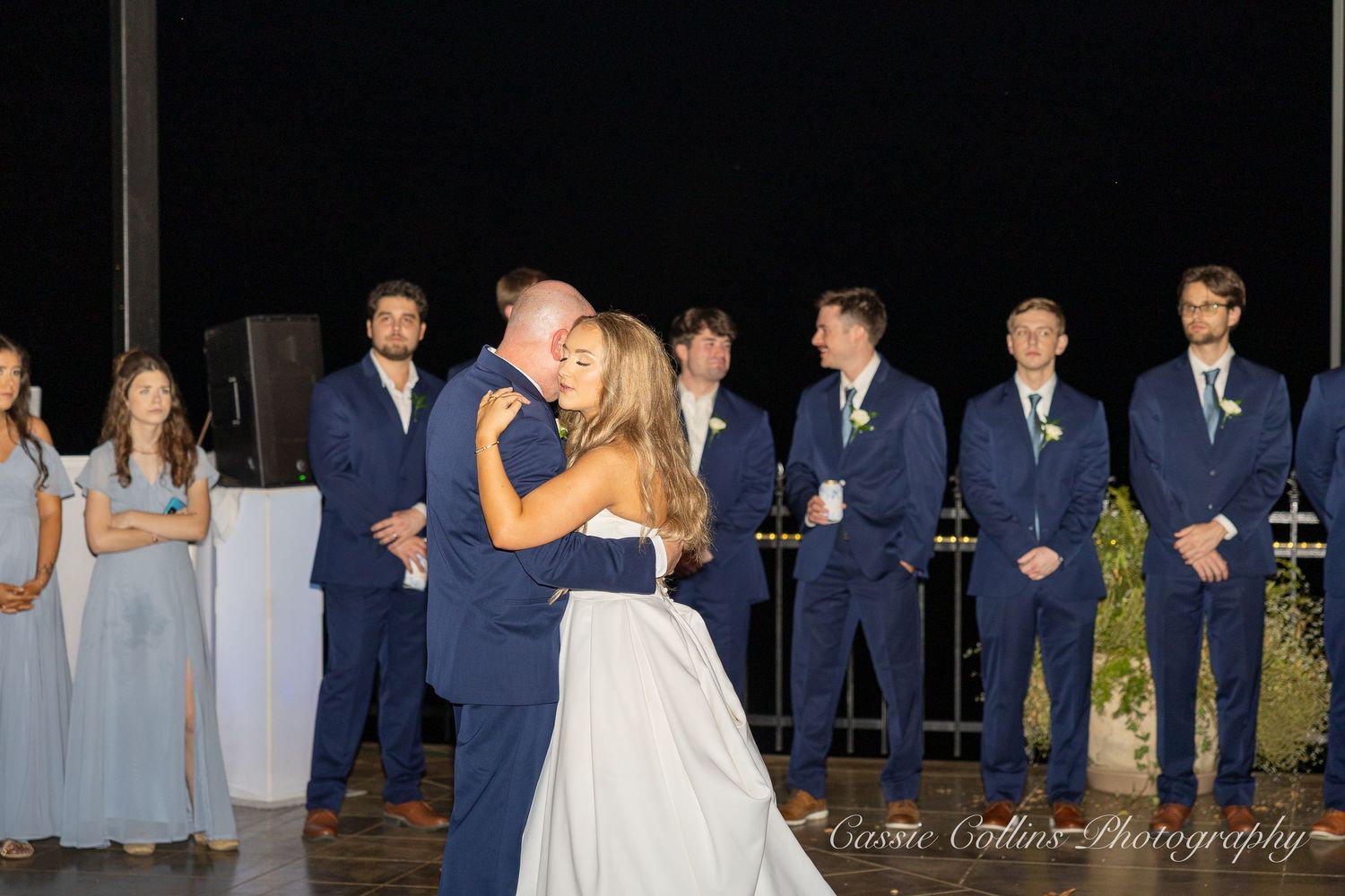 Bride and groom share first dance at outdoor wedding reception while groomsmen in navy suits watch at night.