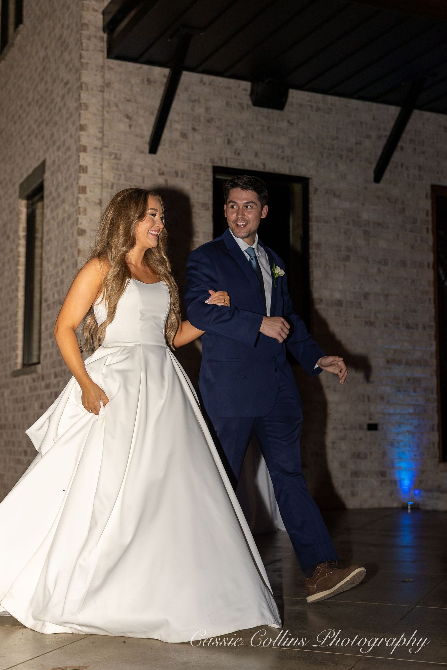 Bride in white gown and groom in navy suit make grand entrance at wedding reception, smiling and dancing together.