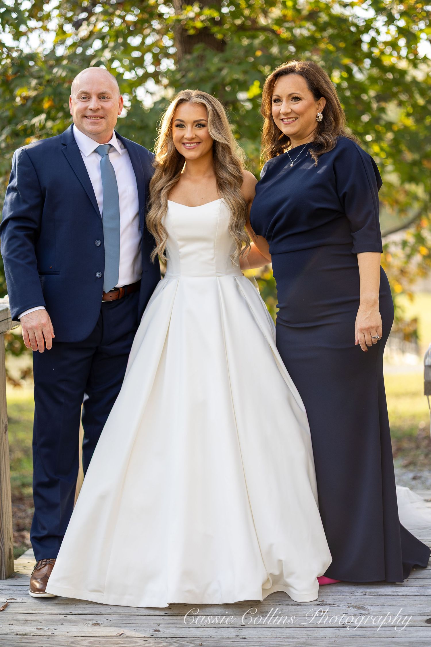 Three people pose outdoors for a formal portrait, with the center figure wearing an elegant white wedding gown.