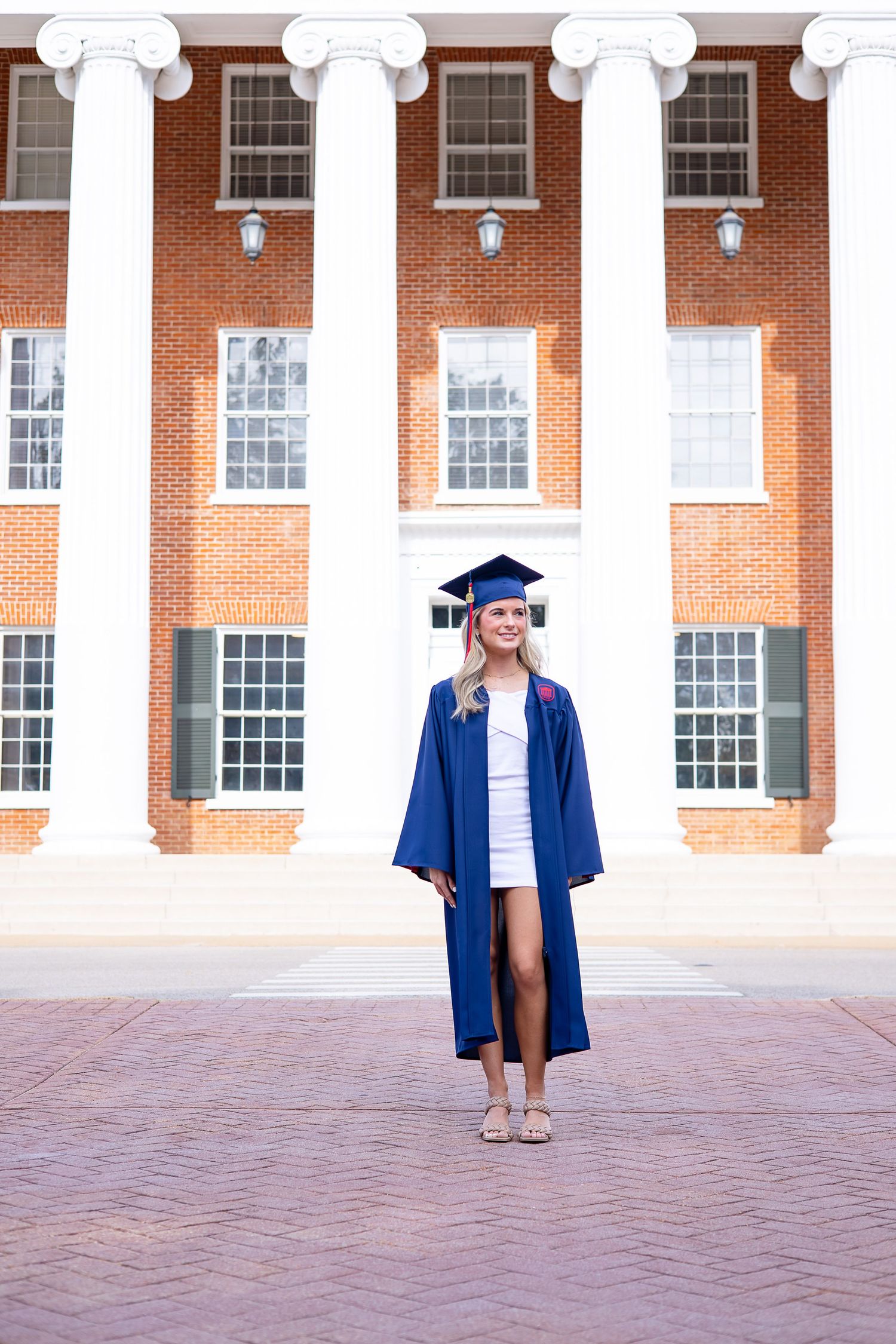 A smiling female graduate poses in blue cap and gown outside a classic brick university building.