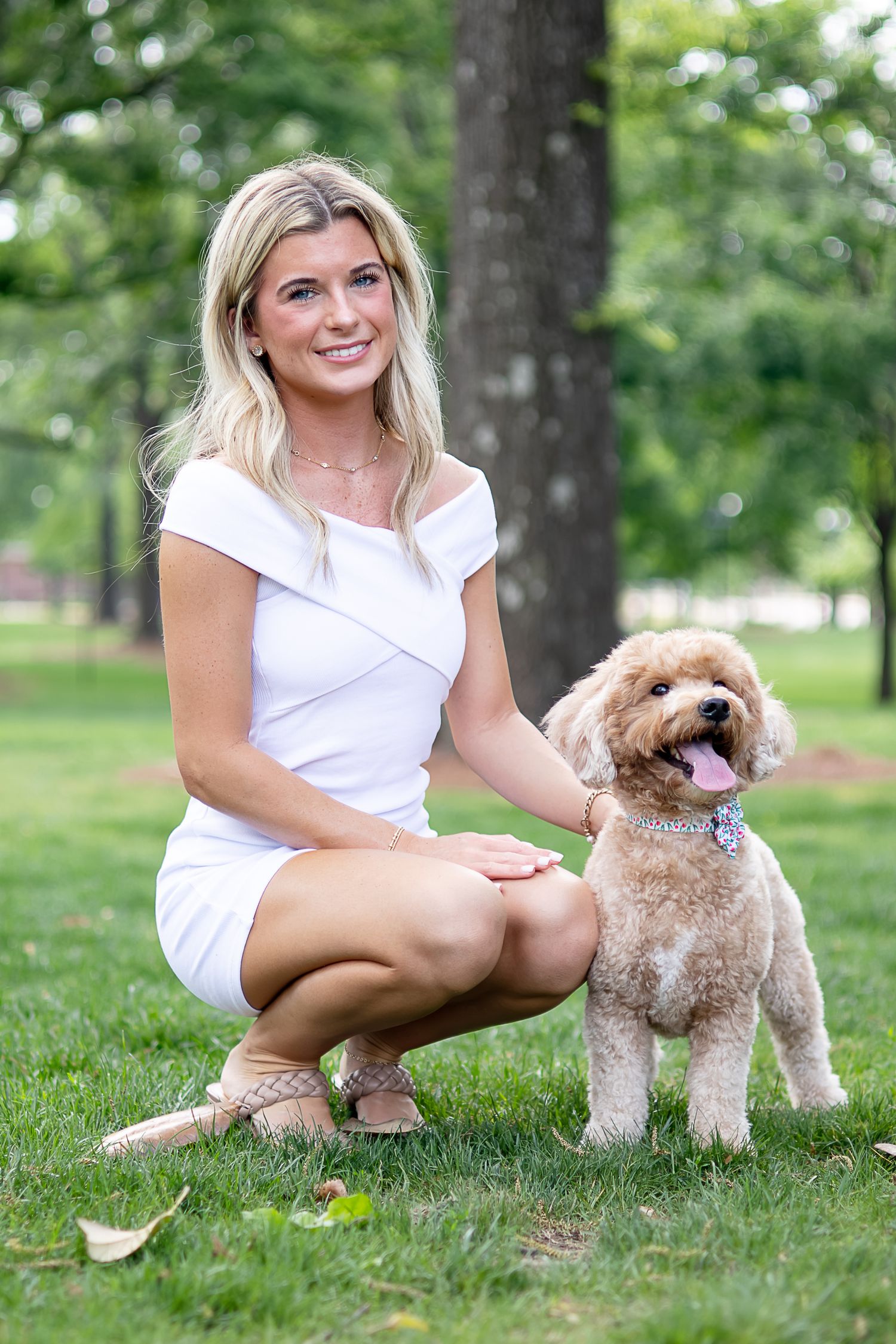 Blonde woman in white dress kneeling on grass in park with fluffy golden doodle dog smiling at camera.