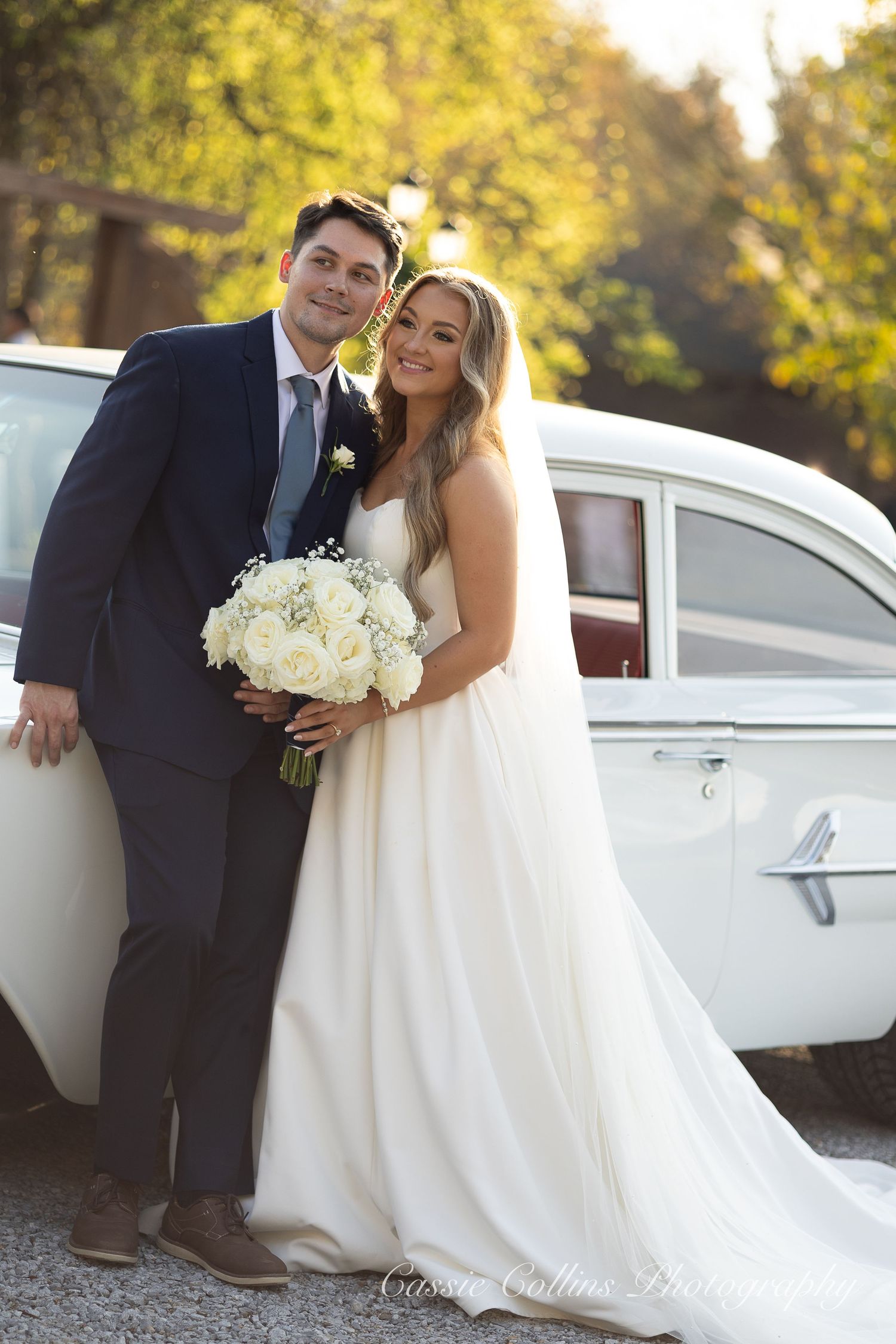 A romantic wedding couple poses with a classic white car in a sunlit autumn setting with golden foliage.