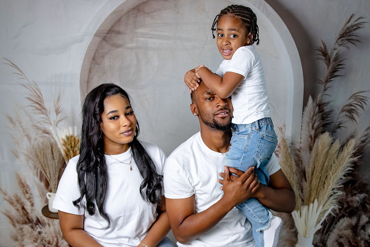 A family of three poses for a studio portrait, parents in white tees with toddler sitting on dad's shoulders.