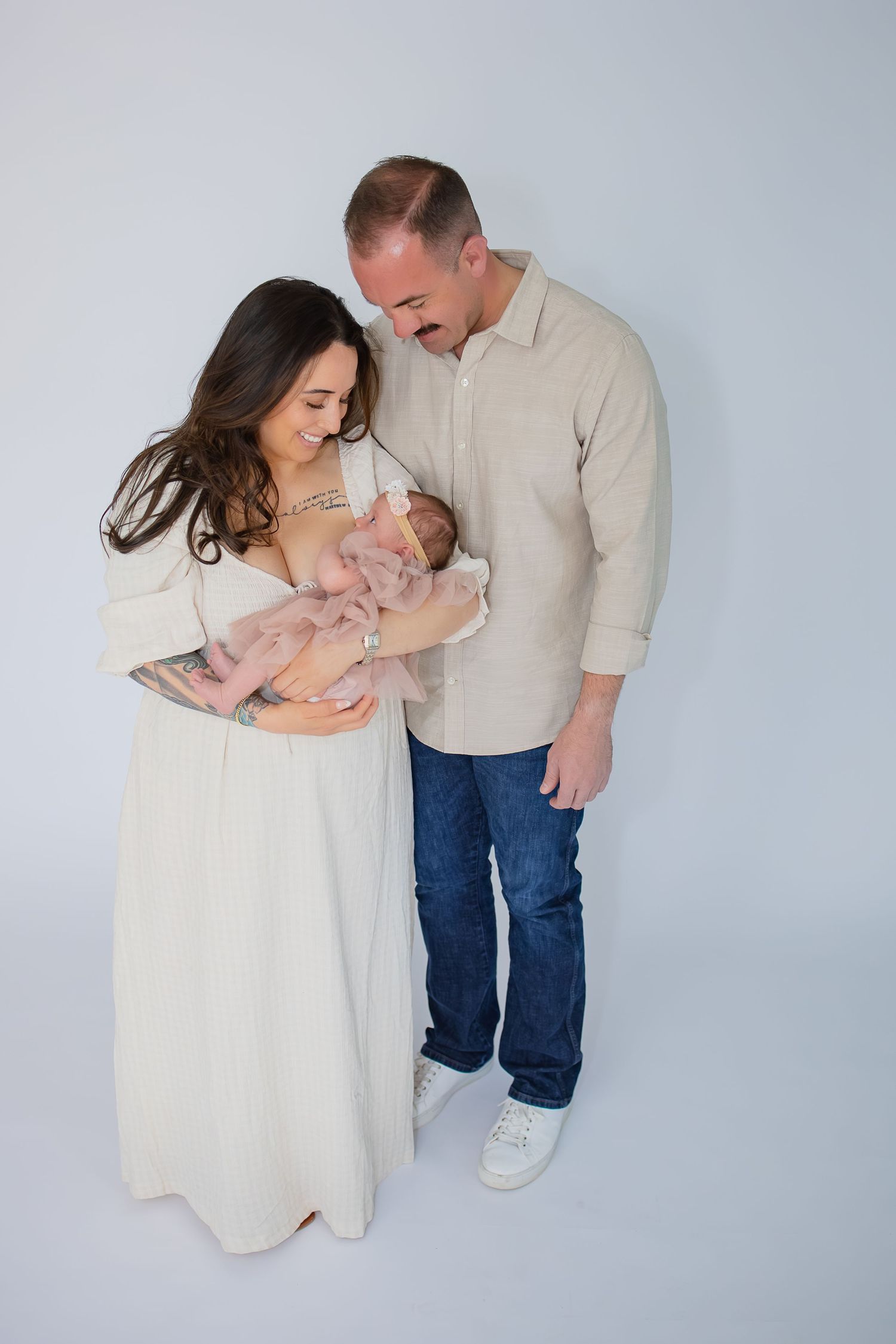 New parents lovingly gaze at their newborn baby in a bright studio portrait, mother in white dress, father in beige shirt.