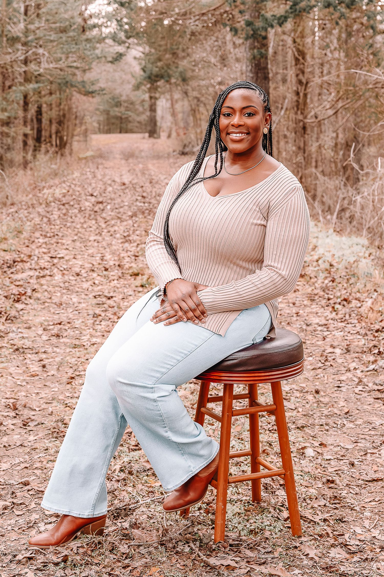 A person in a beige sweater and light jeans sits casually on a wooden stool in a forest setting.