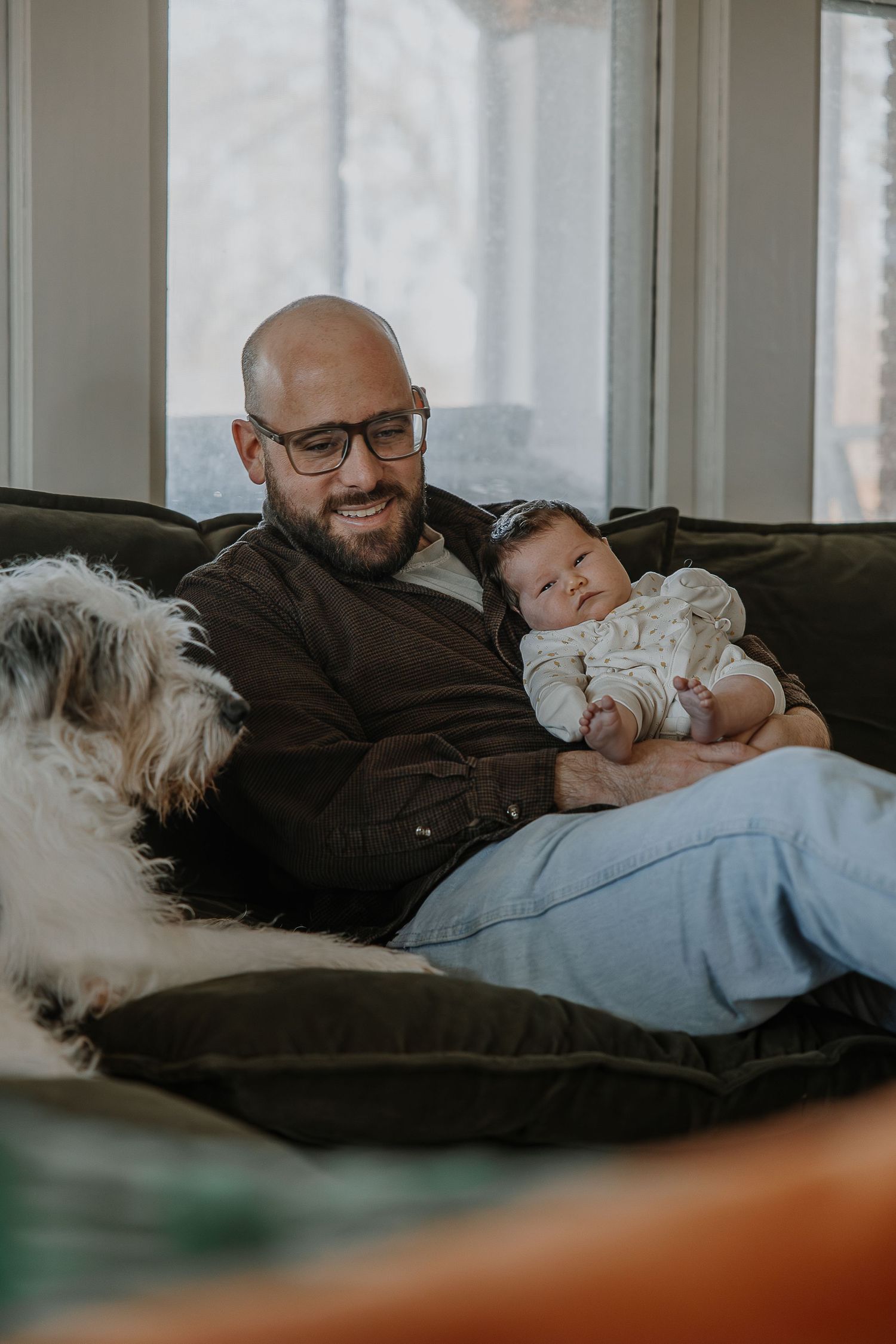 Two people relax together on a couch with a small white dog in a cozy living room setting.