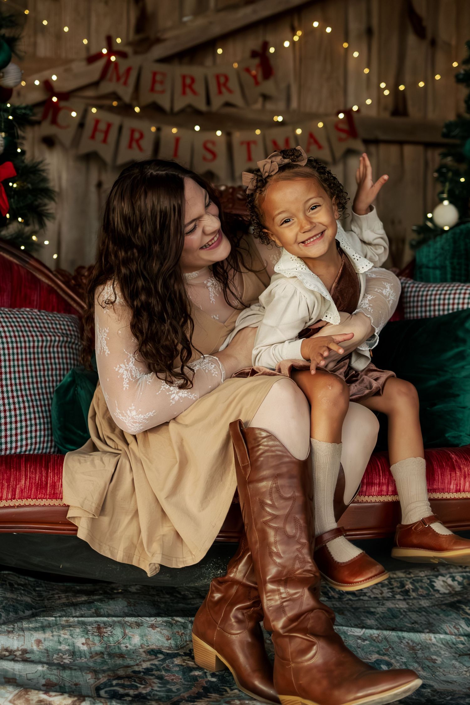 Two friends in cozy winter outfits and brown boots laugh together while sitting on floor with holiday lights in background.