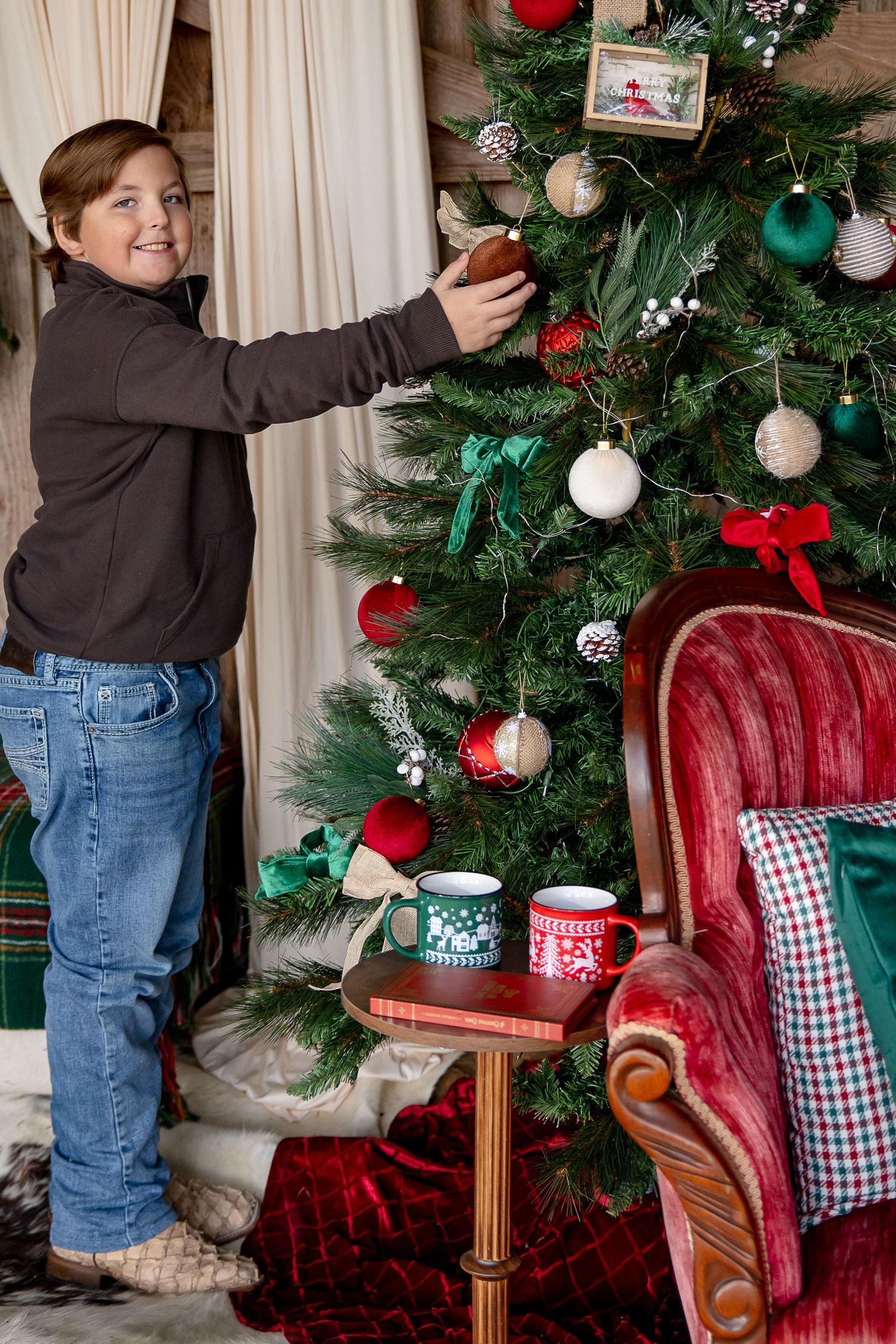 Someone decorates a Christmas tree next to a red velvet chair with festive mugs on a side table.