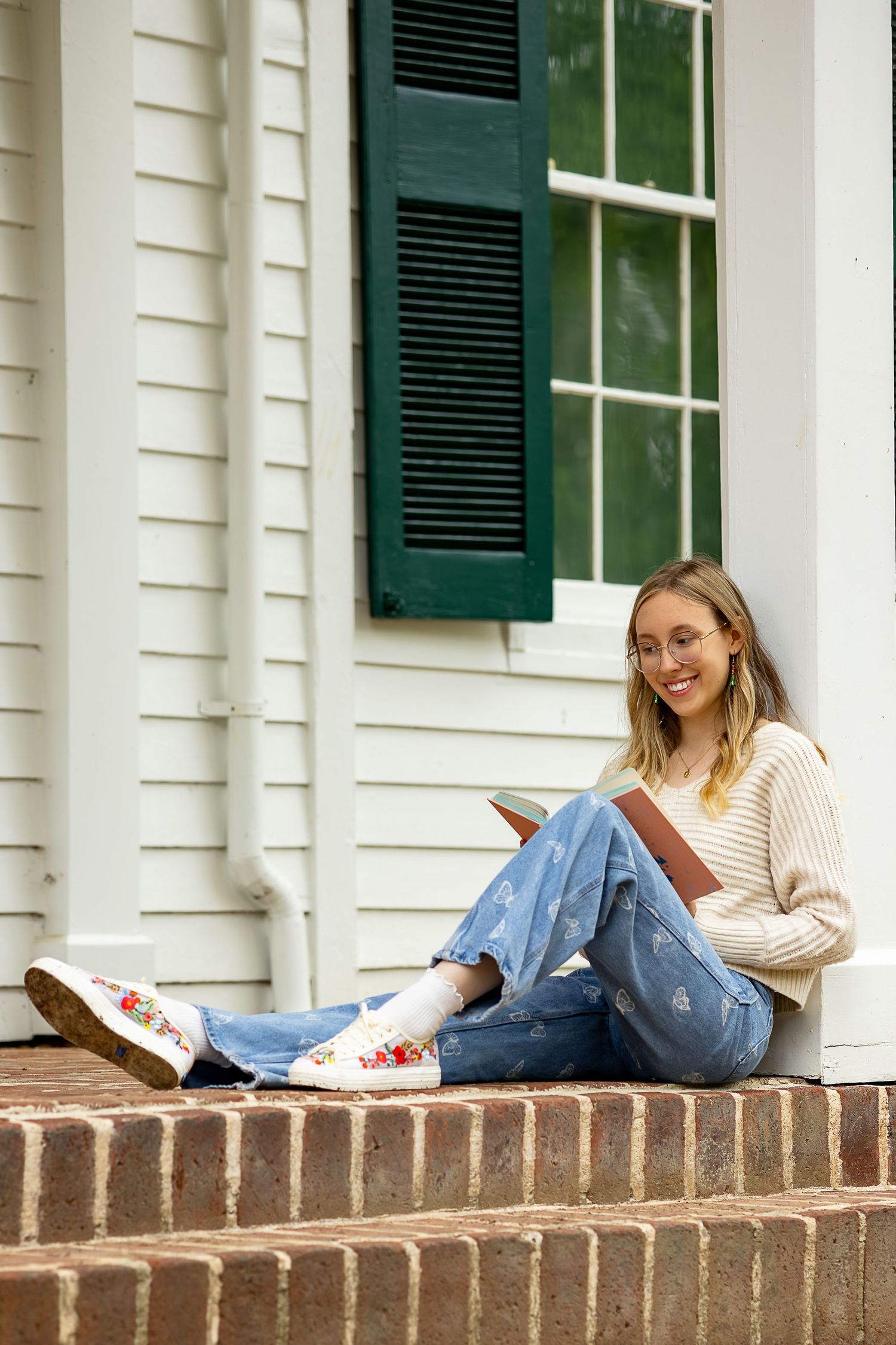 Smiling blonde woman in jeans and cream sweater sitting on brick steps reading a book outside a white house.