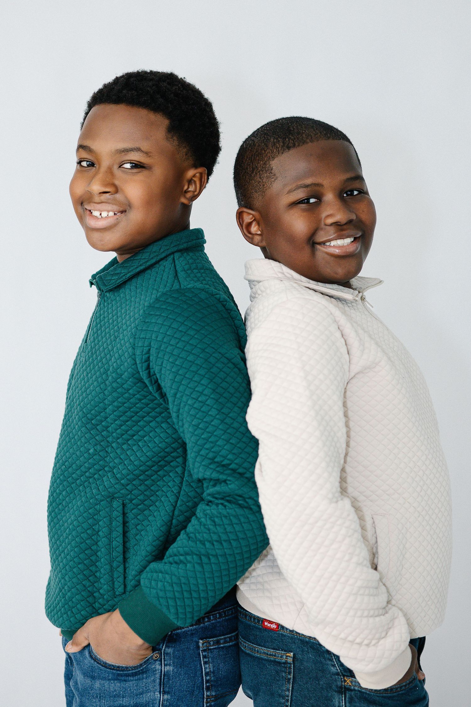 Two young people in quilted sweaters smile while standing back to back against white background.