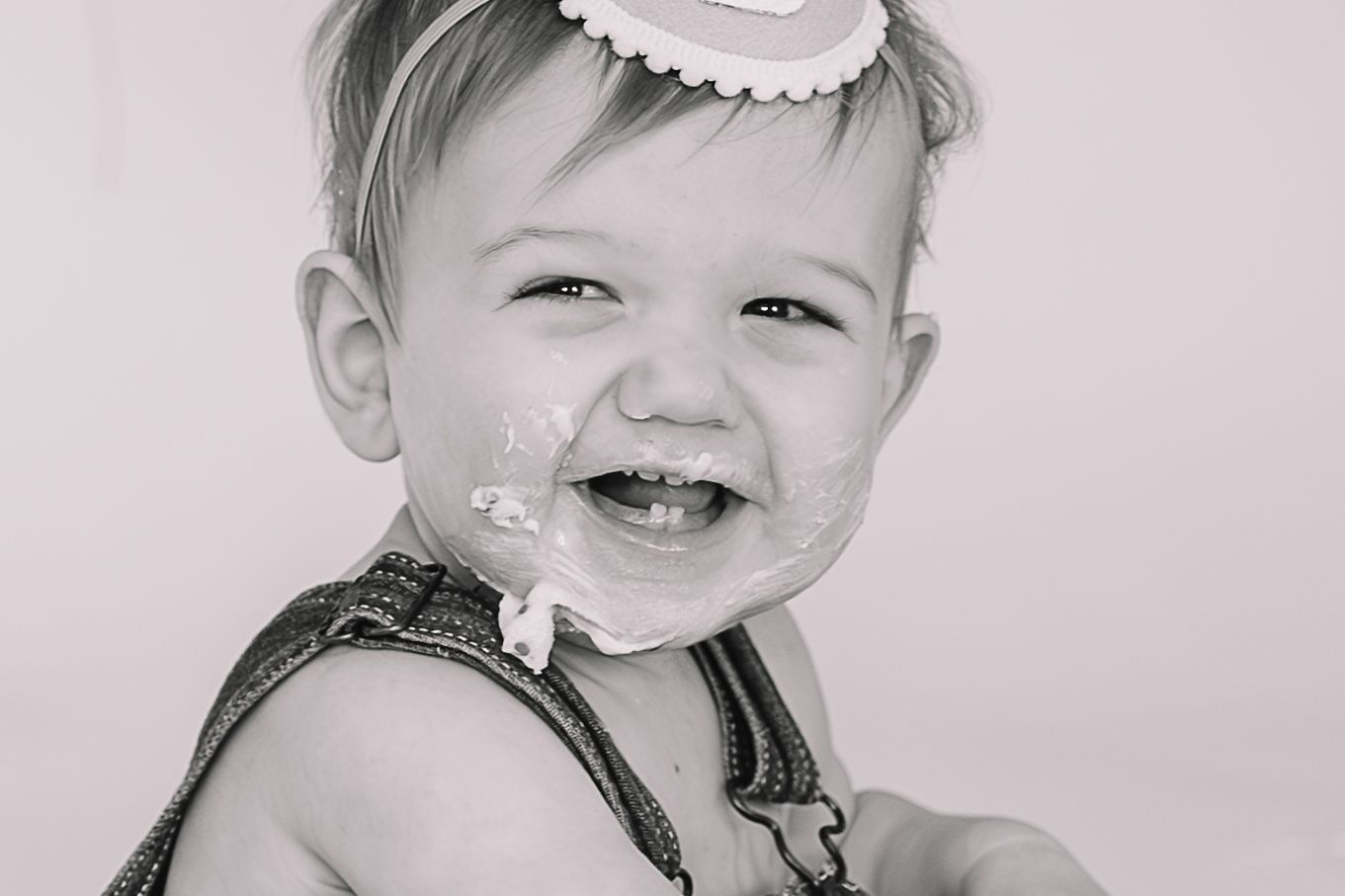 Black and white portrait of a joyful toddler wearing overalls and a white headband.