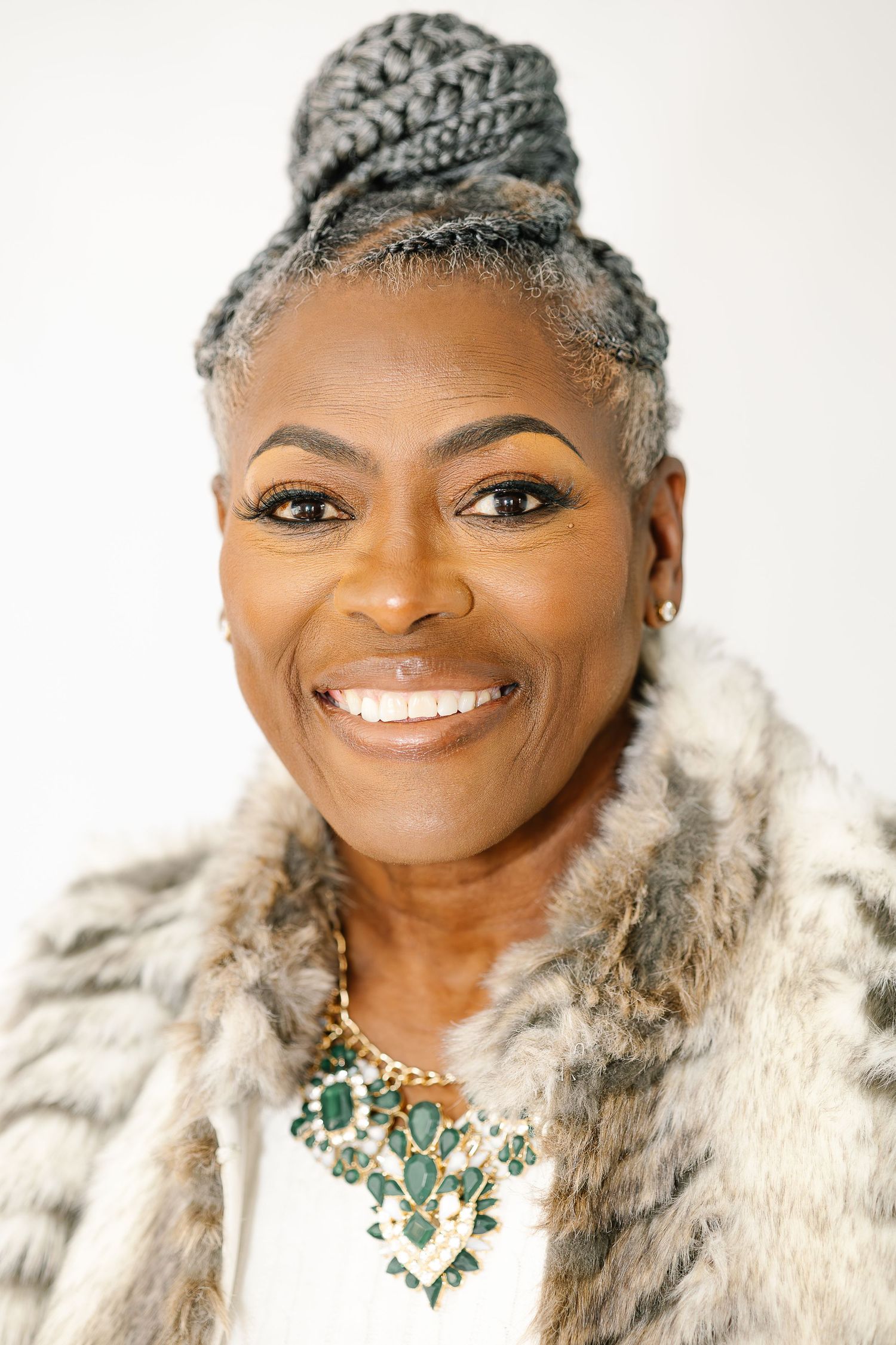A person wearing a fur coat and emerald necklace poses elegantly against a white background.