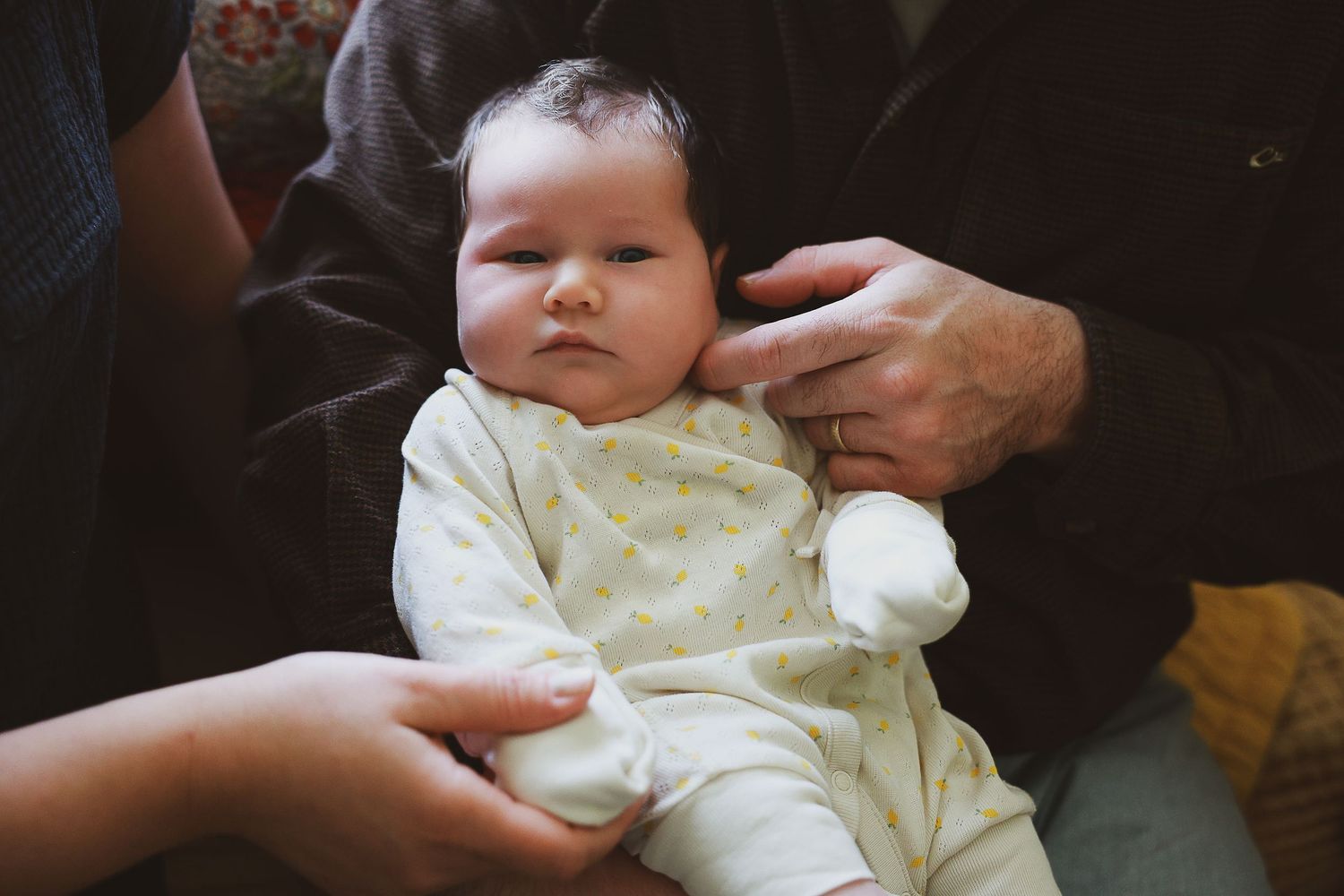 A small baby in a pale yellow outfit is being held by adult hands in a dimly lit setting.