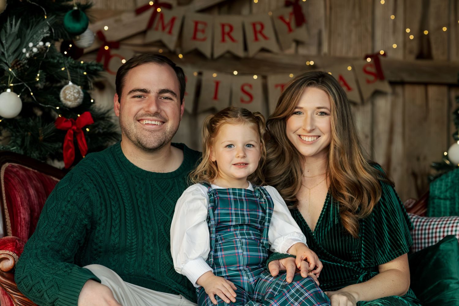 A family poses together by a decorated Christmas tree with a rustic wooden wall and 'Merry' banner in the background.