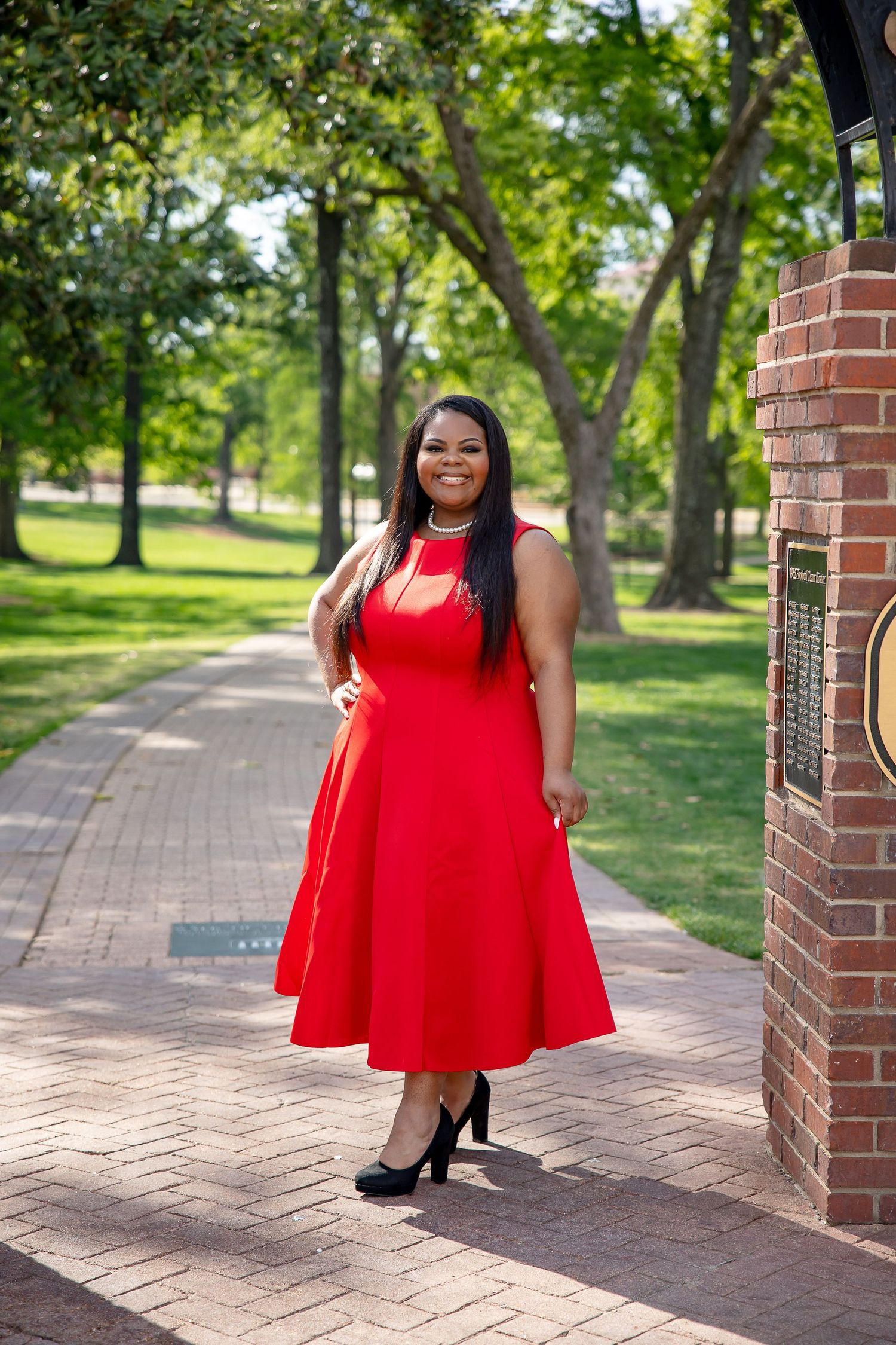 Woman in red midi dress and black heels smiling on brick pathway in lush green park setting.