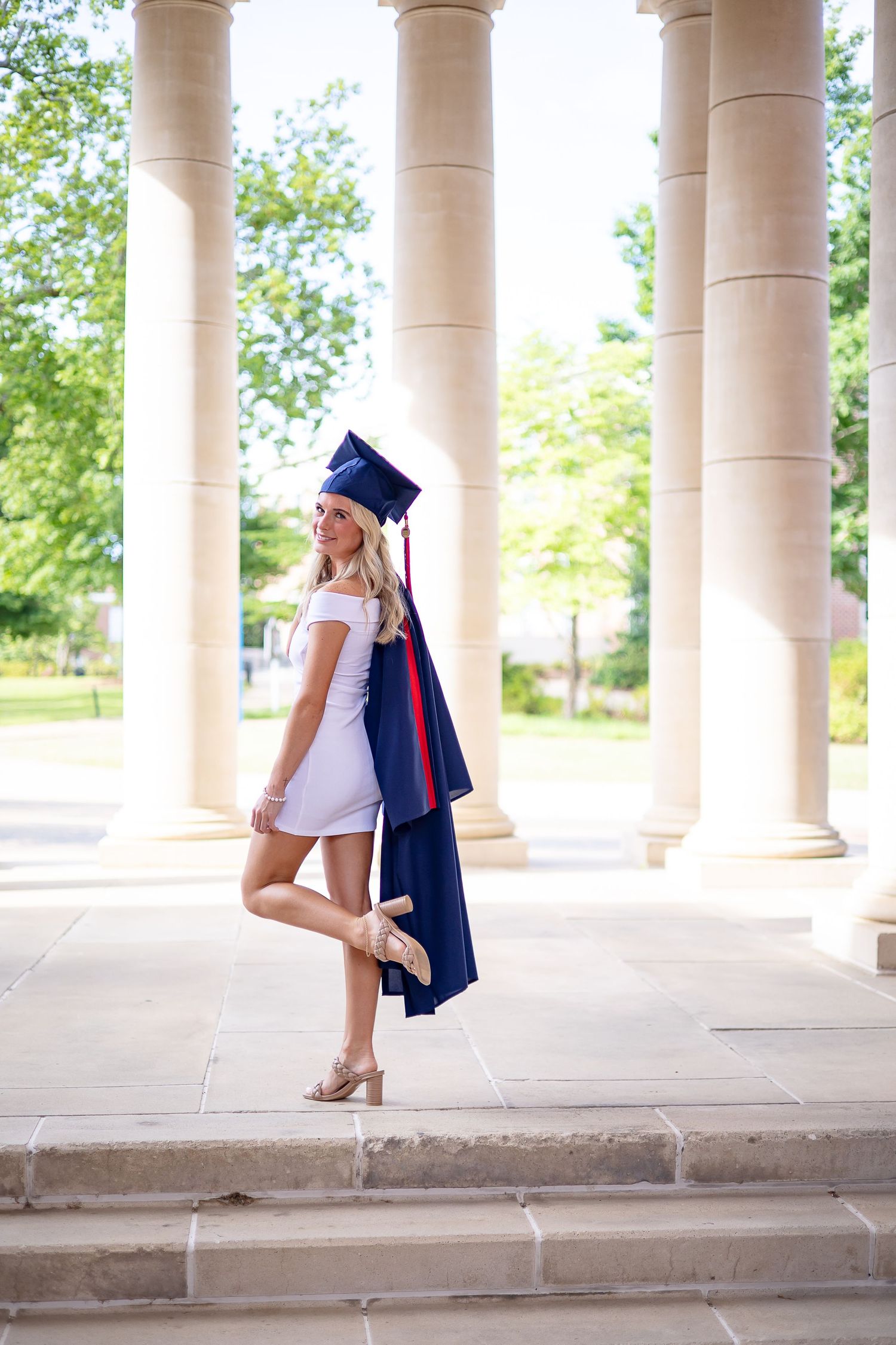 Young woman in white dress and navy graduation cap and gown poses on stone steps between tall columns.