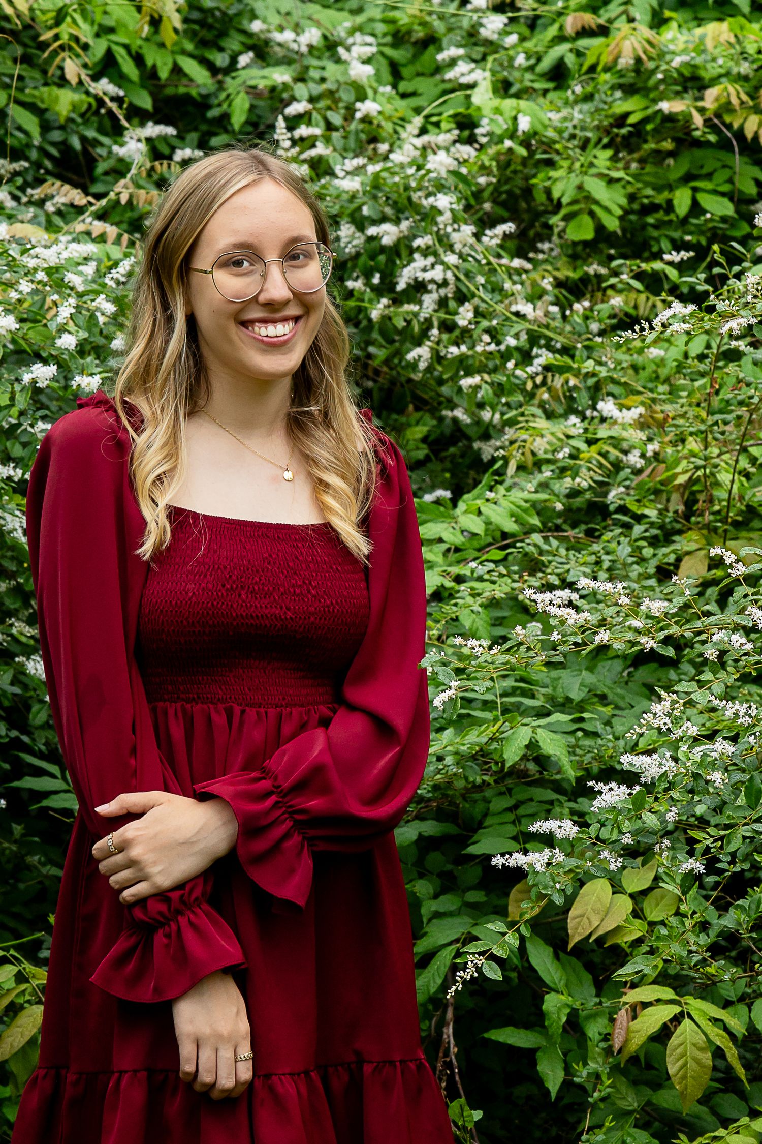 Smiling young woman in red dress and glasses standing in lush green garden with white flowers.