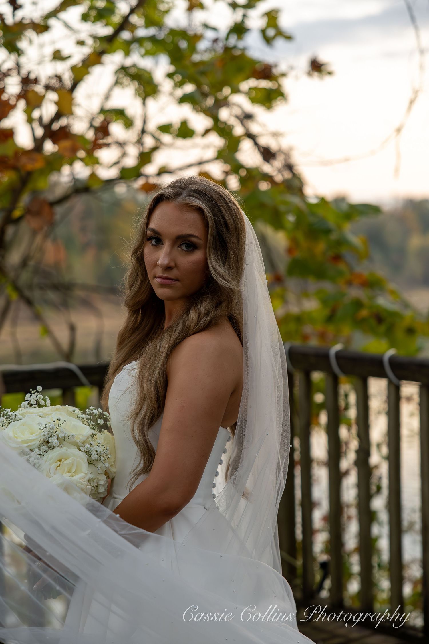 Bride in white wedding dress poses outdoors near water at sunset with autumn foliage in background.